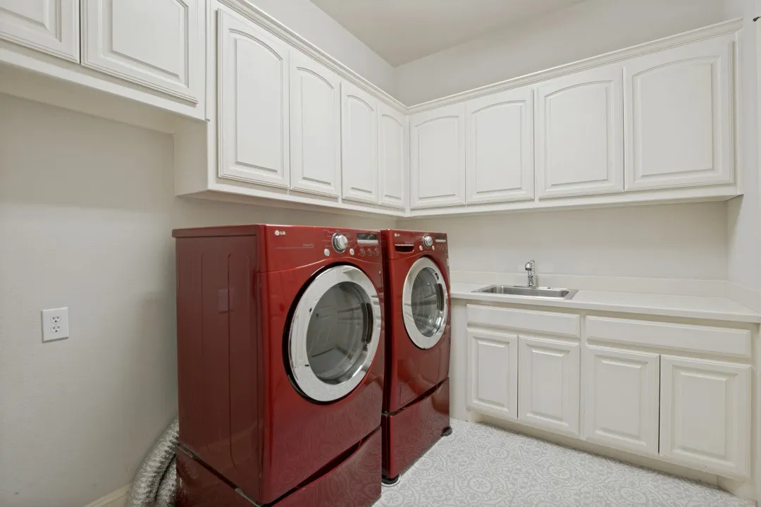 Laundry room featuring extensive white cabinetry, a built-in sink with a chrome faucet, light-toned countertops, and patterned floor tiling