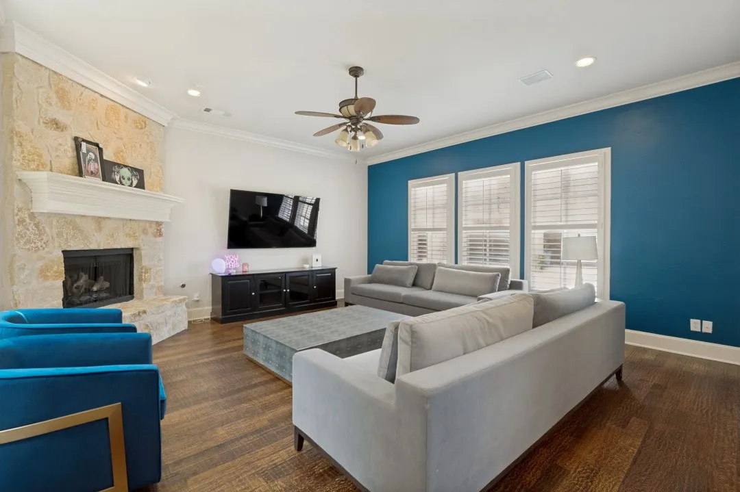 Spacious living area featuring a stone-clad fireplace with a white mantel, rich wood-finish flooring, and multiple windows with plantation shutters