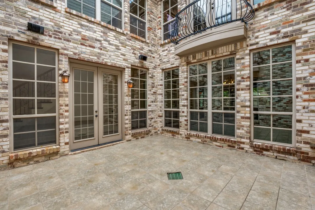 Paved patio area featuring multi-tone brick exterior, multiple large windows with grid patterns, double French doors, and a decorative wrought iron balcony