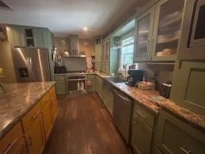 Kitchen featuring wood-finish flooring, two-tone cabinetry with glass-front uppers, stainless steel appliances, a built-in range hood, and stone countertops