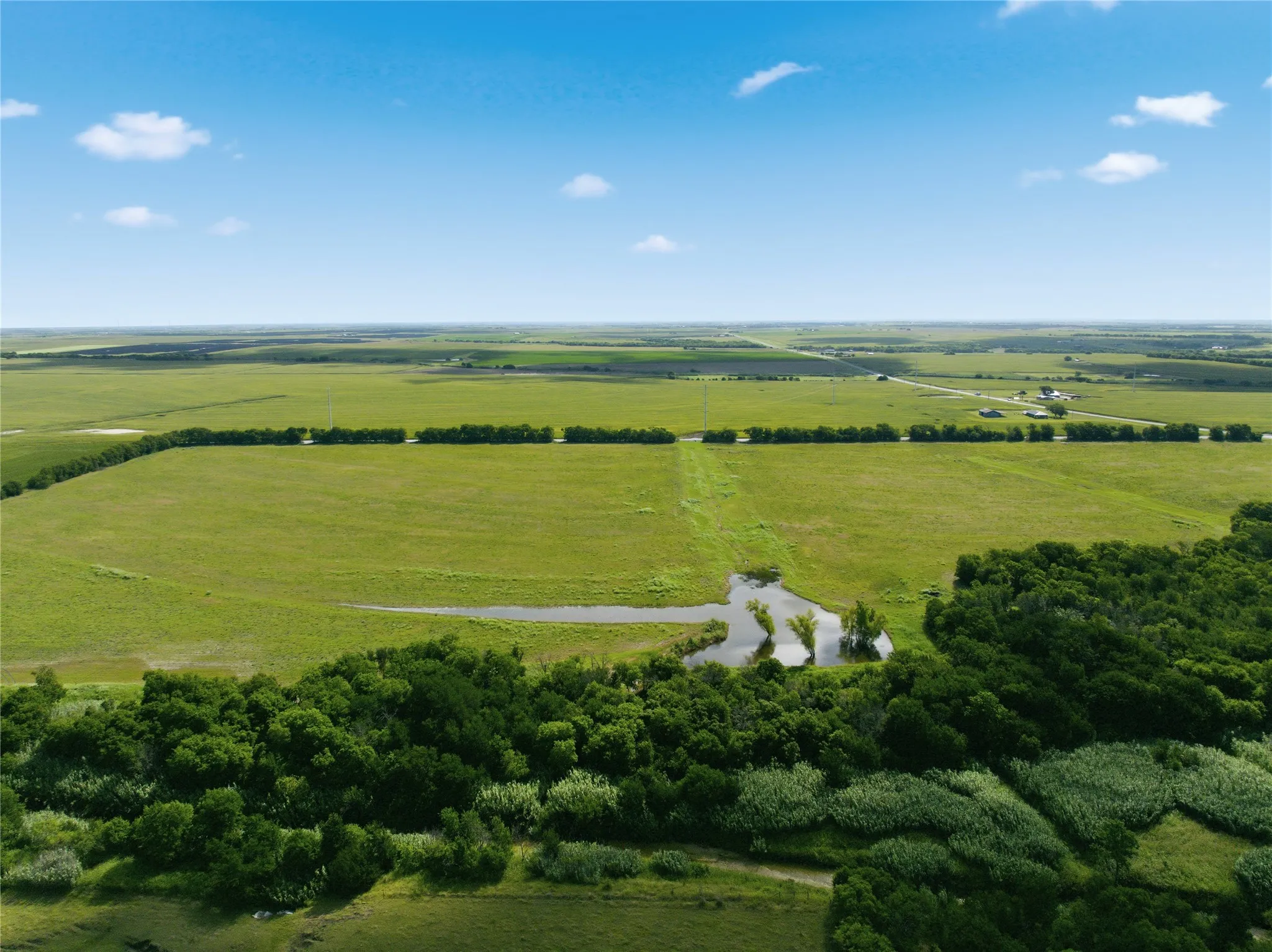 Expansive green fields under a clear blue sky, featuring a distant rural road and scattered buildings