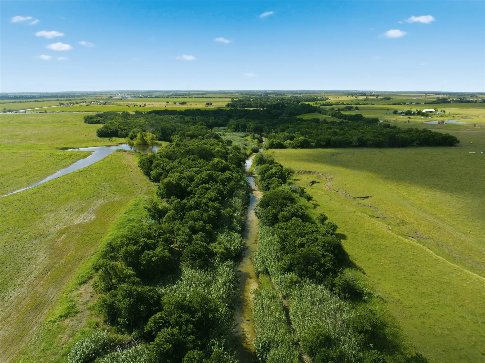 Expansive green landscape featuring a winding waterway, natural tree lines, open fields, and distant structures under a clear sky