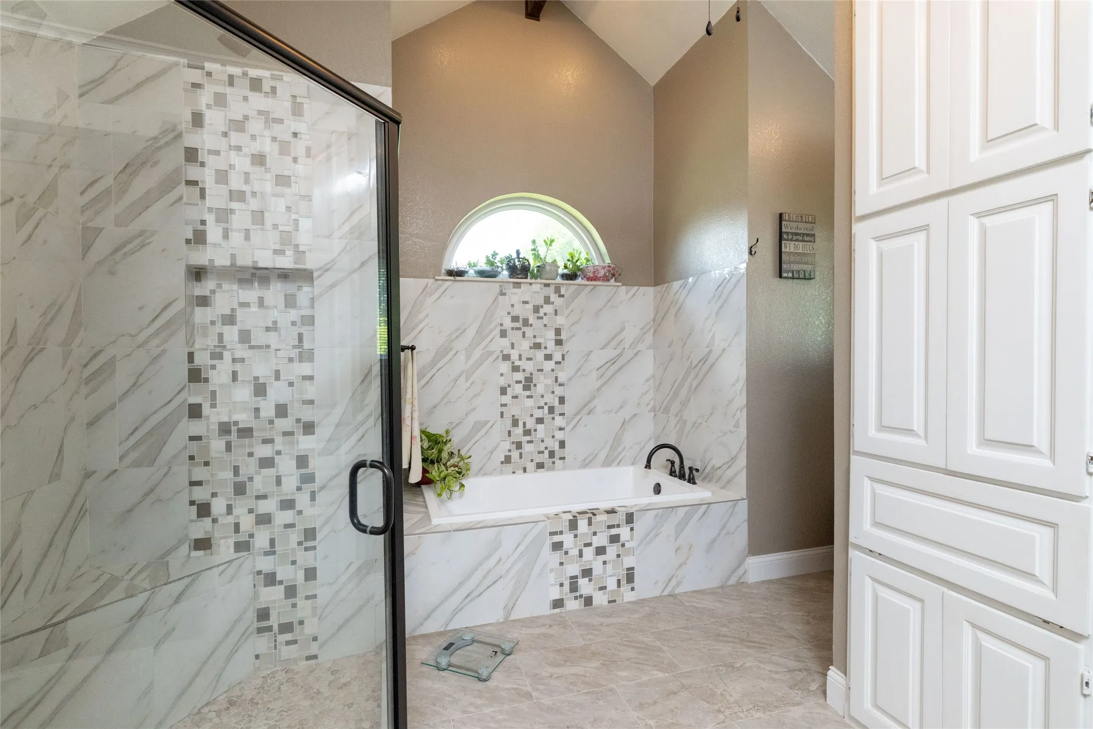 Bathroom featuring a walk-in shower with glass enclosure, built-in soaking tub, arched window, and extensive white cabinetry