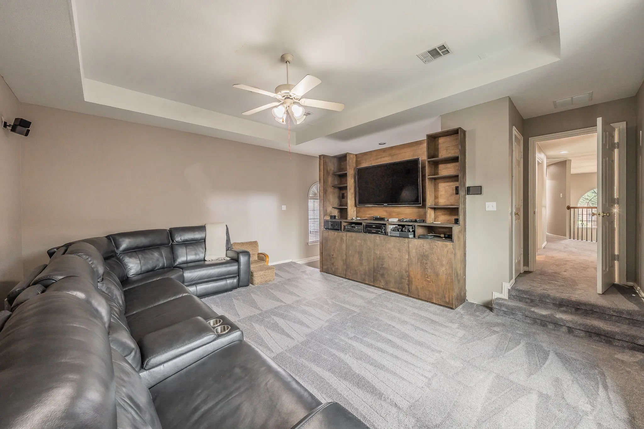 Spacious media room featuring a built-in entertainment center with open shelving, tray ceiling with a ceiling fan, and patterned carpeting