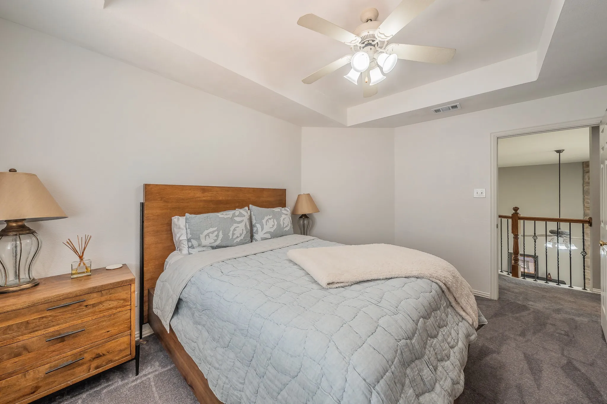 Recessed tray ceiling with ceiling fan, wood-finish headboard, and gray carpet flooring