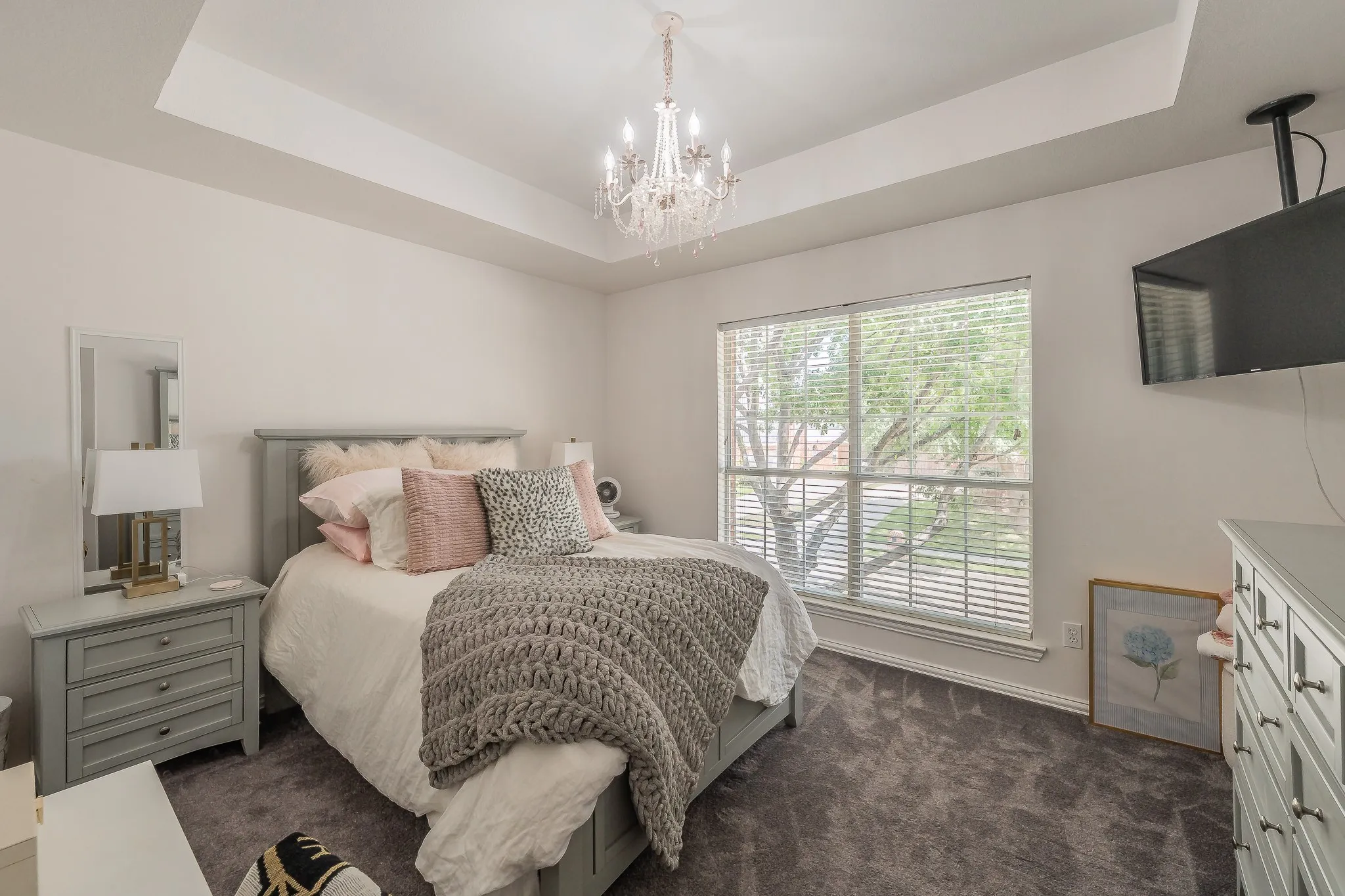 Spacious room featuring a tray ceiling with an ornate chandelier, a large window with horizontal blinds, and dark gray carpet flooring