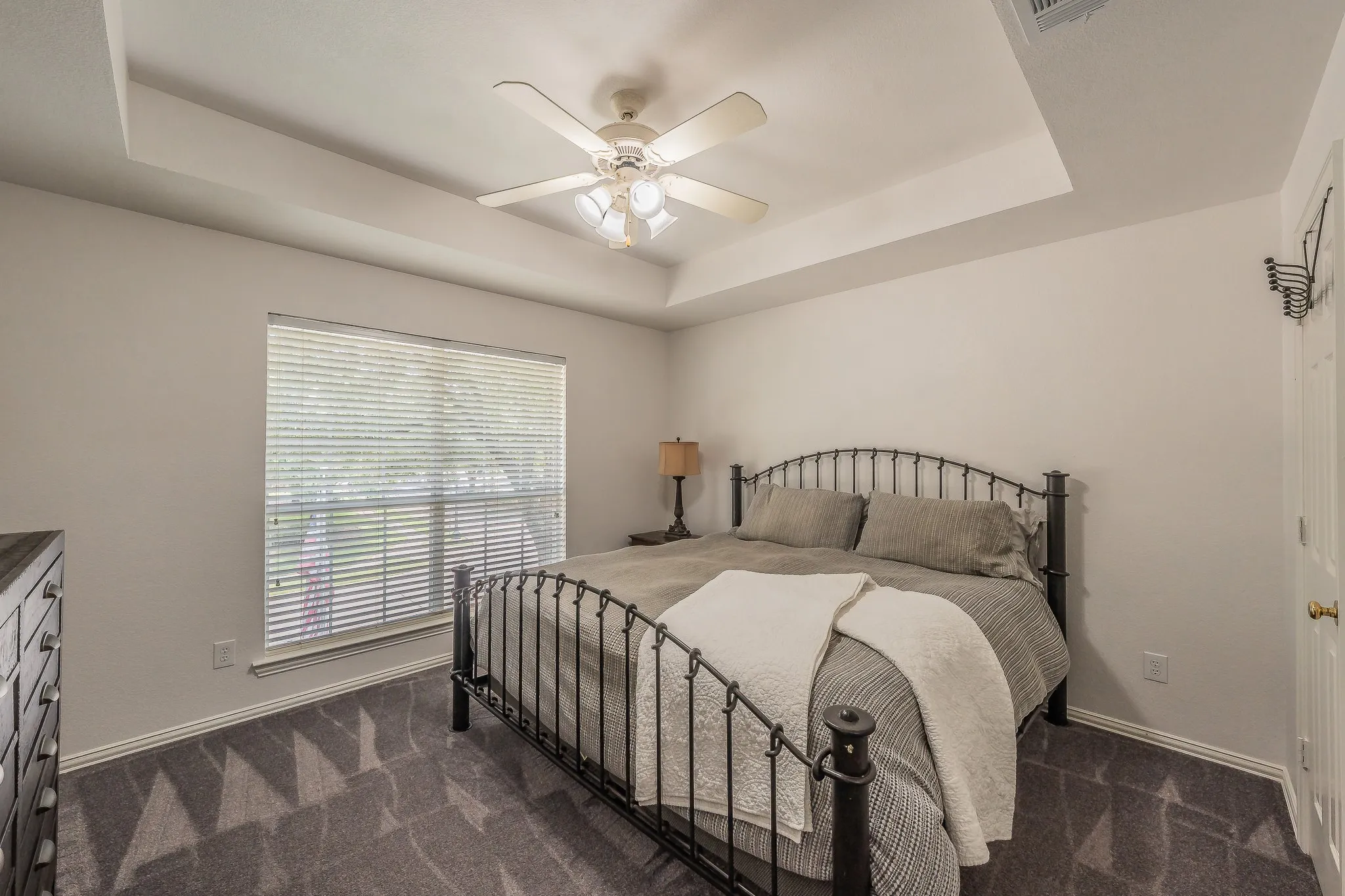 Recessed tray ceiling with a ceiling fan, large window with blinds, and patterned carpet flooring