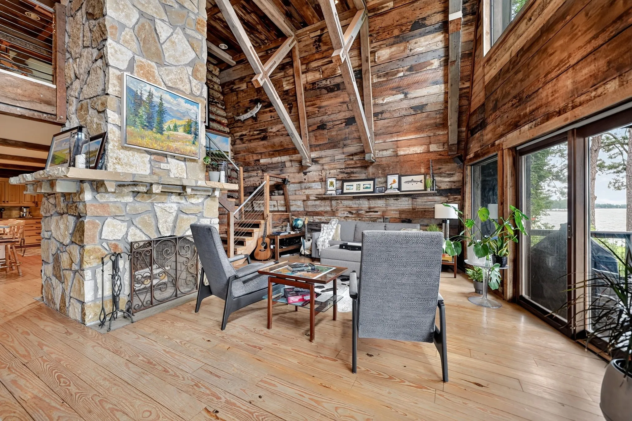 Stone-clad fireplace with mantel, hardwood flooring, wood-paneled walls, vaulted wood-beam ceiling, and large sliding glass doors opening to outdoor space