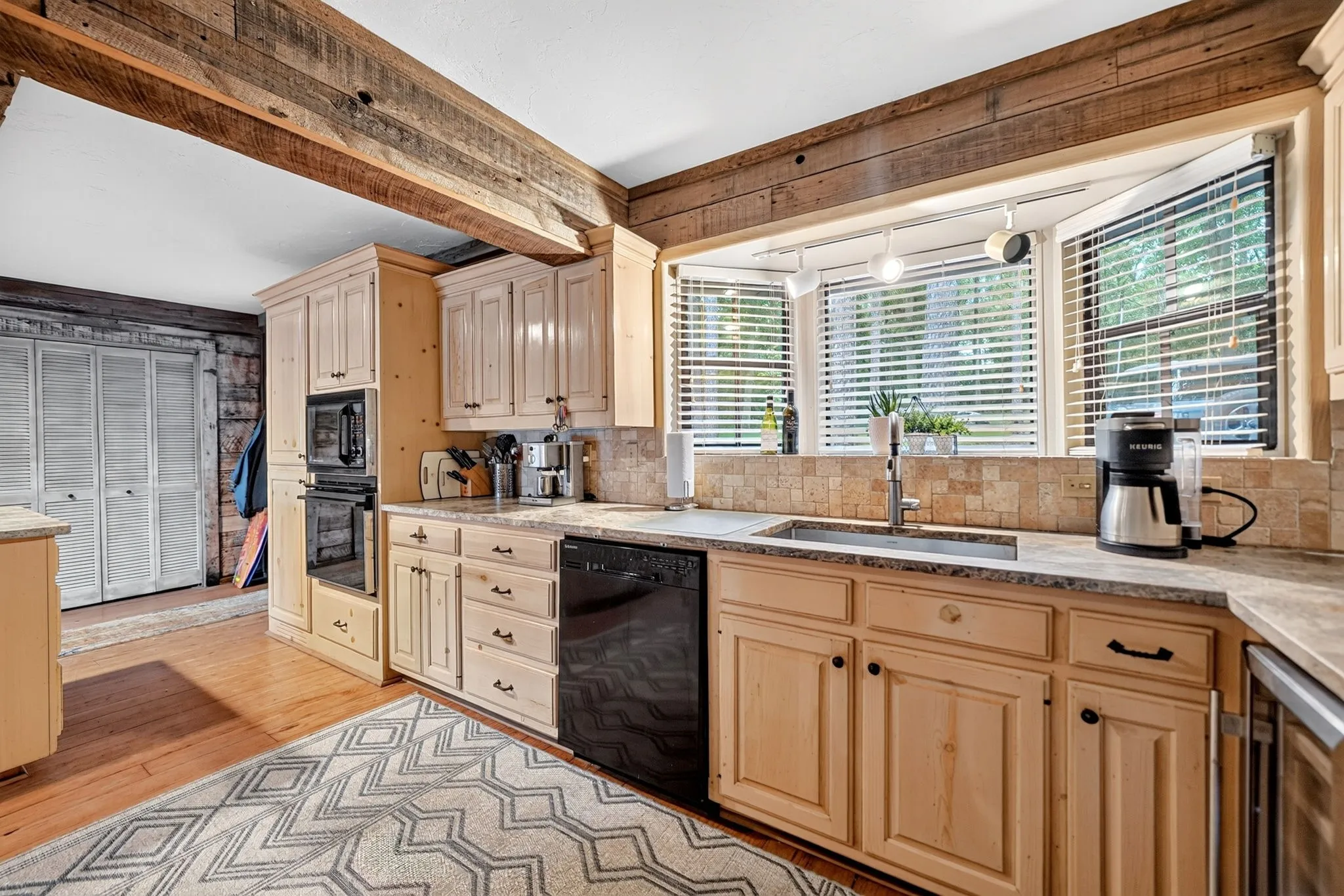 Kitchen featuring a bay window, light wood cabinetry, stone-finish countertops, black dishwasher, and built-in wall oven