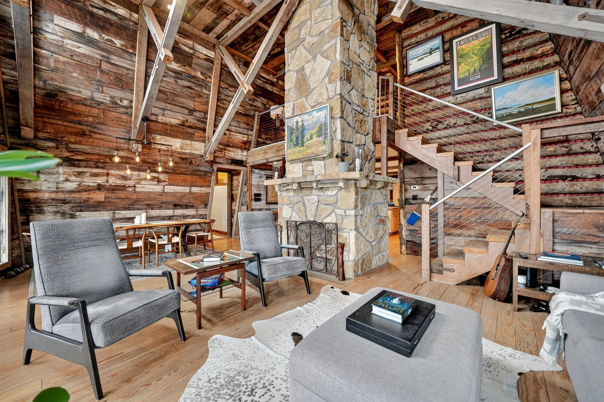 Expansive great room featuring a stone fireplace, exposed timber beams, wood-finish flooring, a contemporary staircase with cable railing, and a dining area with a linear pendant light fixture