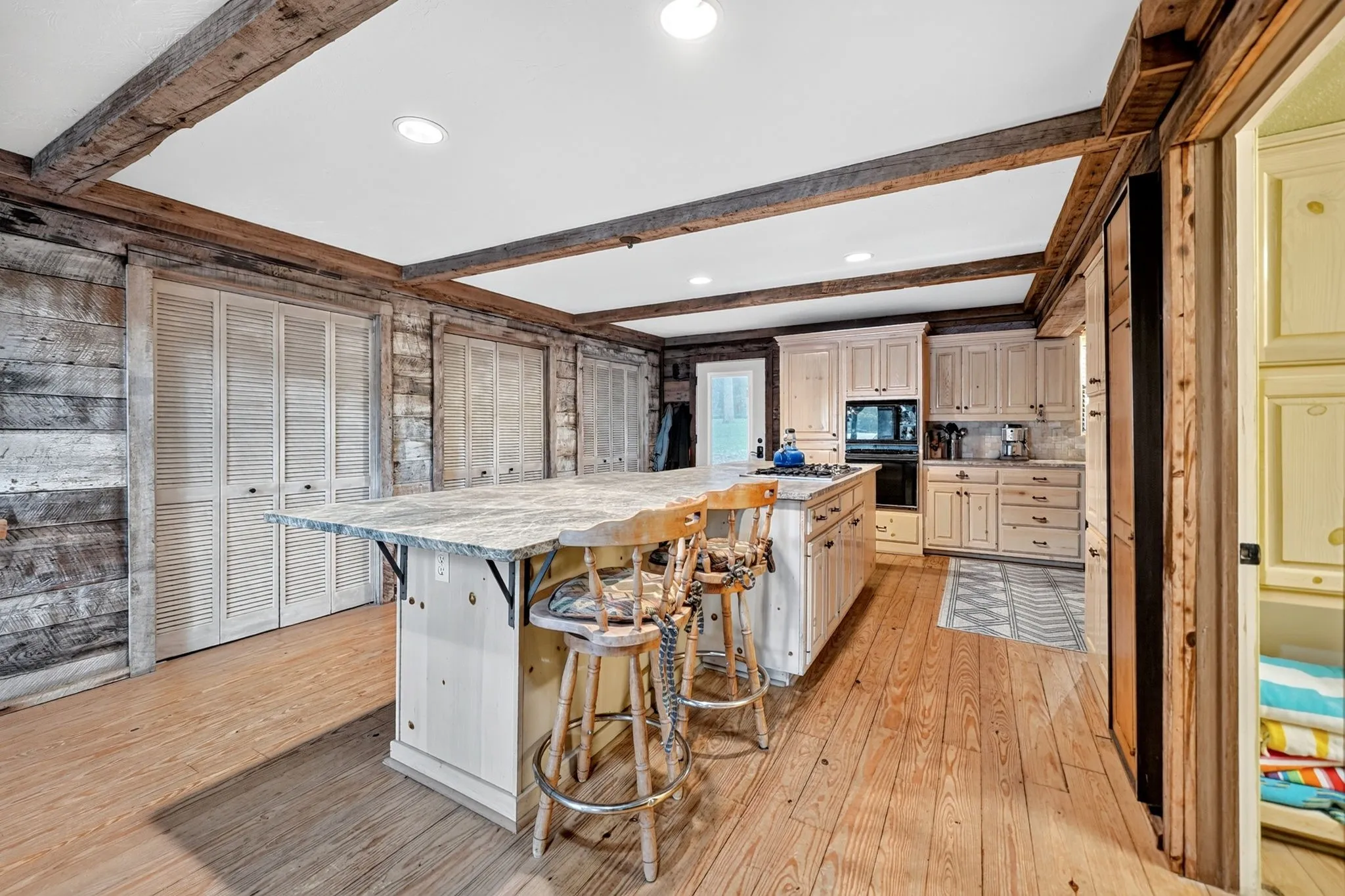 Kitchen featuring a large central island with a natural stone countertop, exposed wood ceiling beams, wood-finish flooring, light-toned cabinetry, and integrated appliances