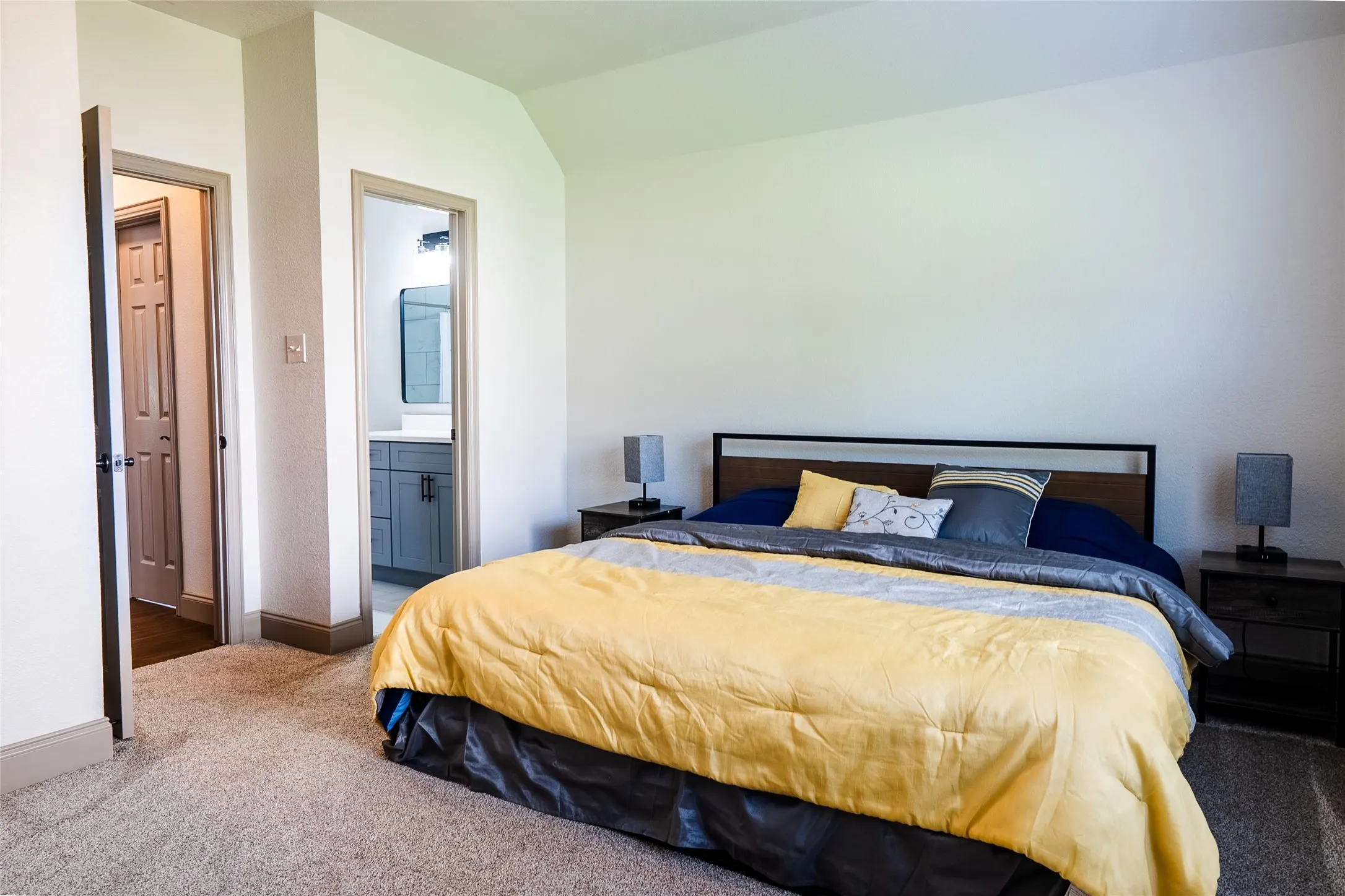 Carpeted bedroom featuring a vaulted ceiling and direct access to an attached bathroom with a single vanity and rectangular mirror