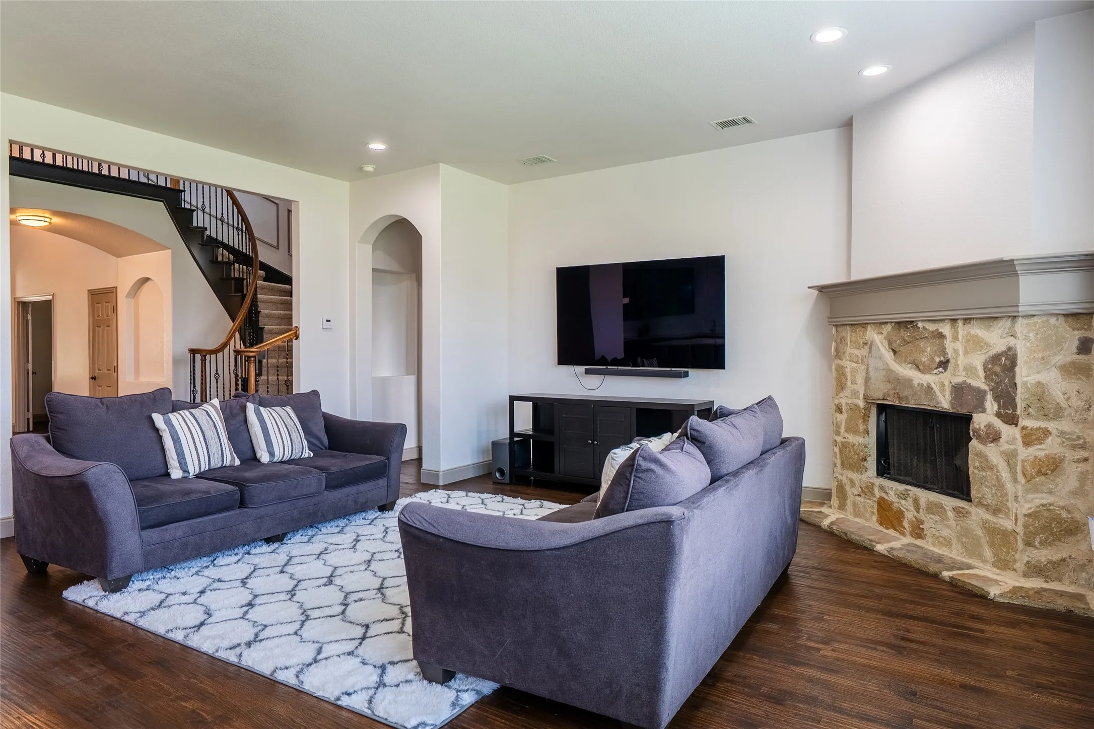 Spacious living area featuring rich wood-finish flooring, a stone fireplace with a substantial mantel, and white walls