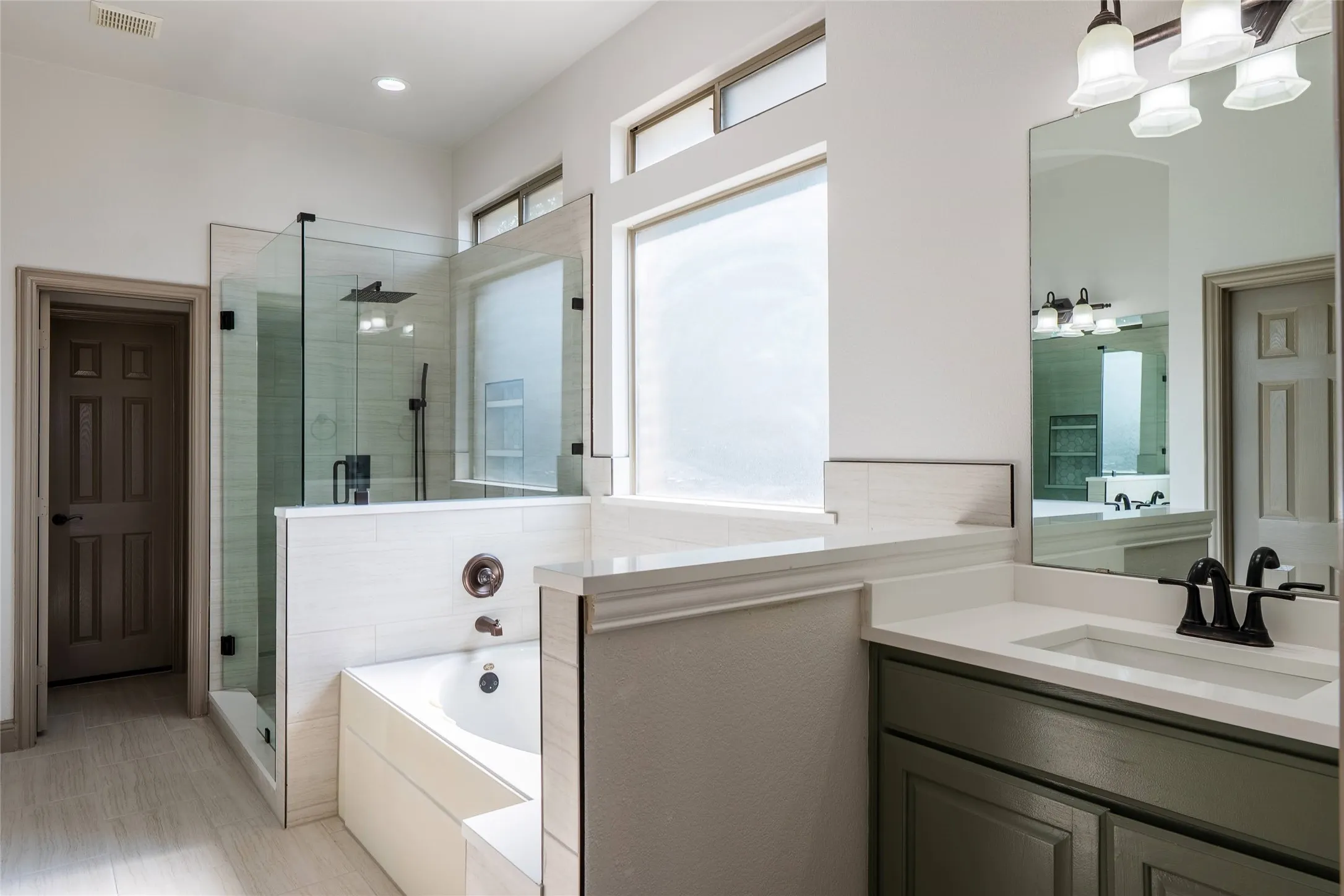 Contemporary bathroom featuring a built-in tub, a glass-enclosed shower with a rainfall showerhead, a vanity with a white countertop and dark green cabinetry, and transom windows for natural illumination