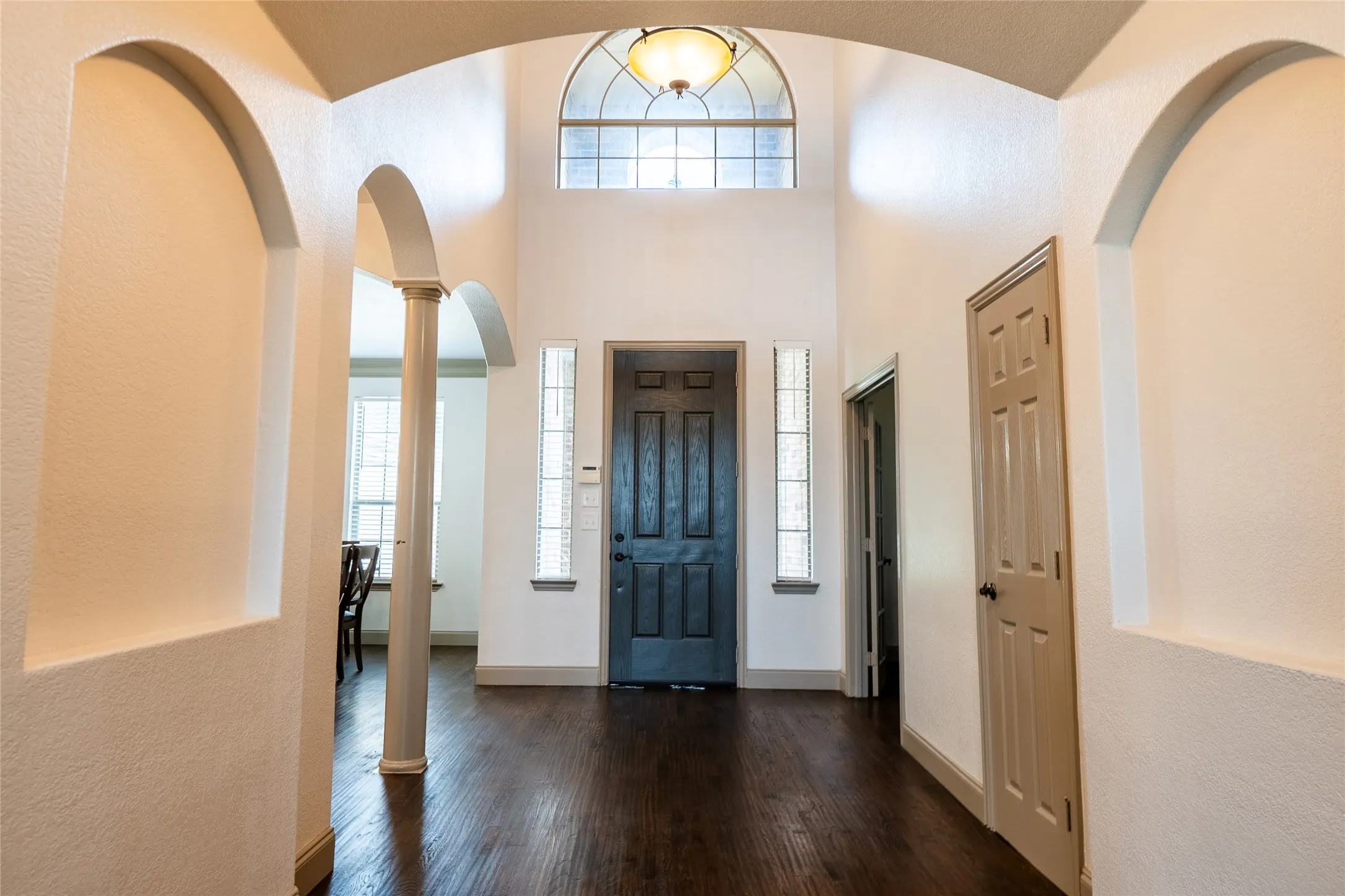 Two-story foyer featuring dark wood-finish flooring, arched doorways, a decorative column, and a transom window above the entry door