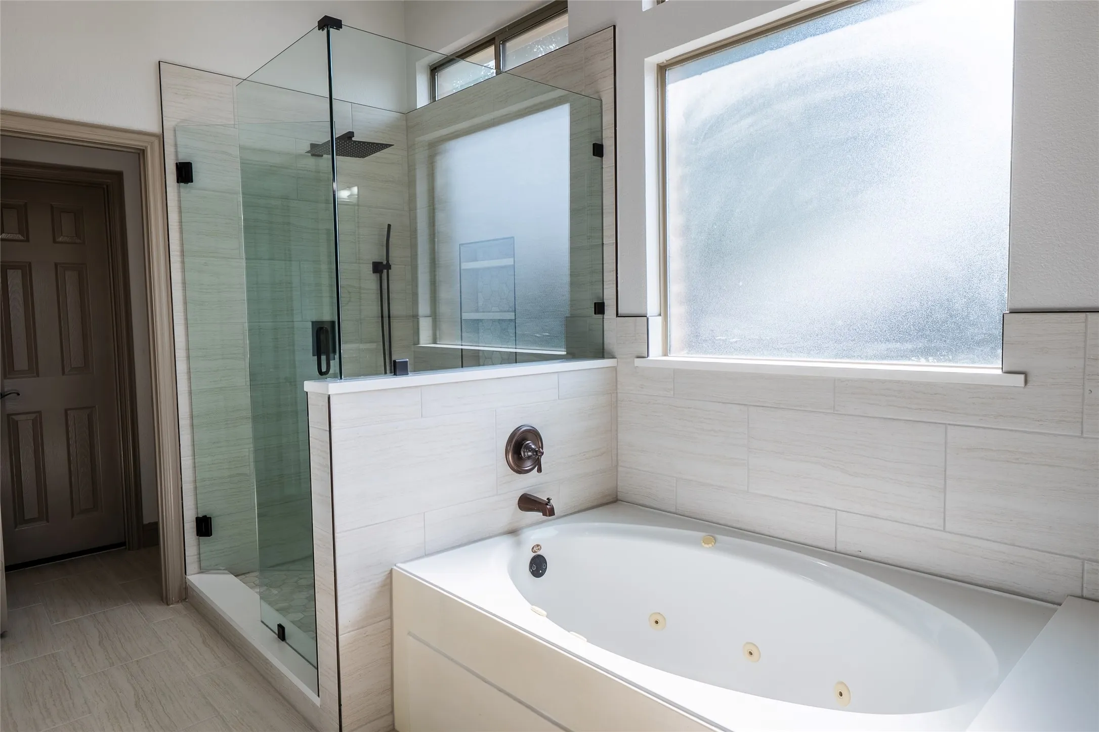 Bathroom featuring a soaking tub with jets, a glass-enclosed shower with dark bronze fixtures, and light-toned tile flooring