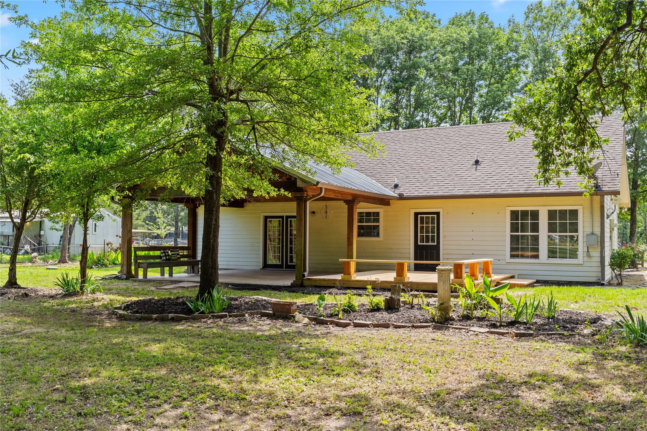 Expansive rear exterior featuring a covered patio with exposed wood beams, a spacious deck, mature trees, and landscaping beds