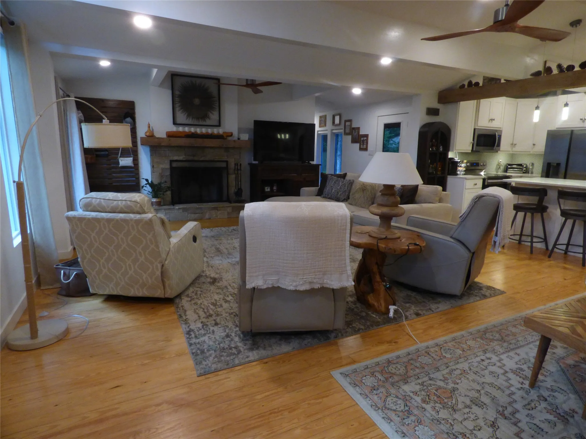 Open-concept living area featuring wood-finish flooring, a stone fireplace with a wood mantle, recessed lighting, and a kitchen with white cabinetry and an island with bar seating