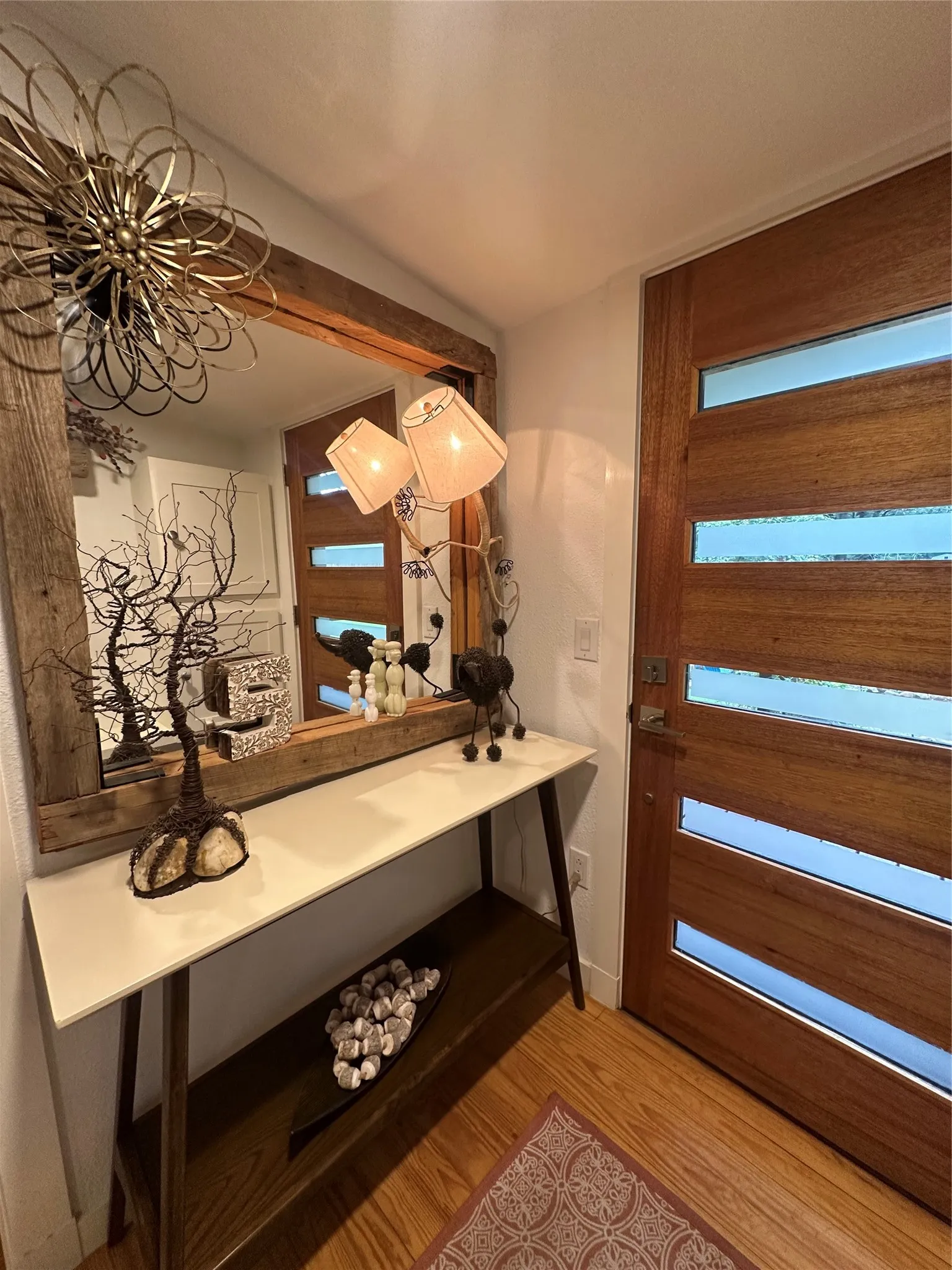 Entryway featuring a solid wood door with horizontal frosted glass insets, wood-finish flooring, a wall-mounted mirror with a rustic wood frame, a console table with a light-toned top, and a ceiling-mounted decorative light fixture