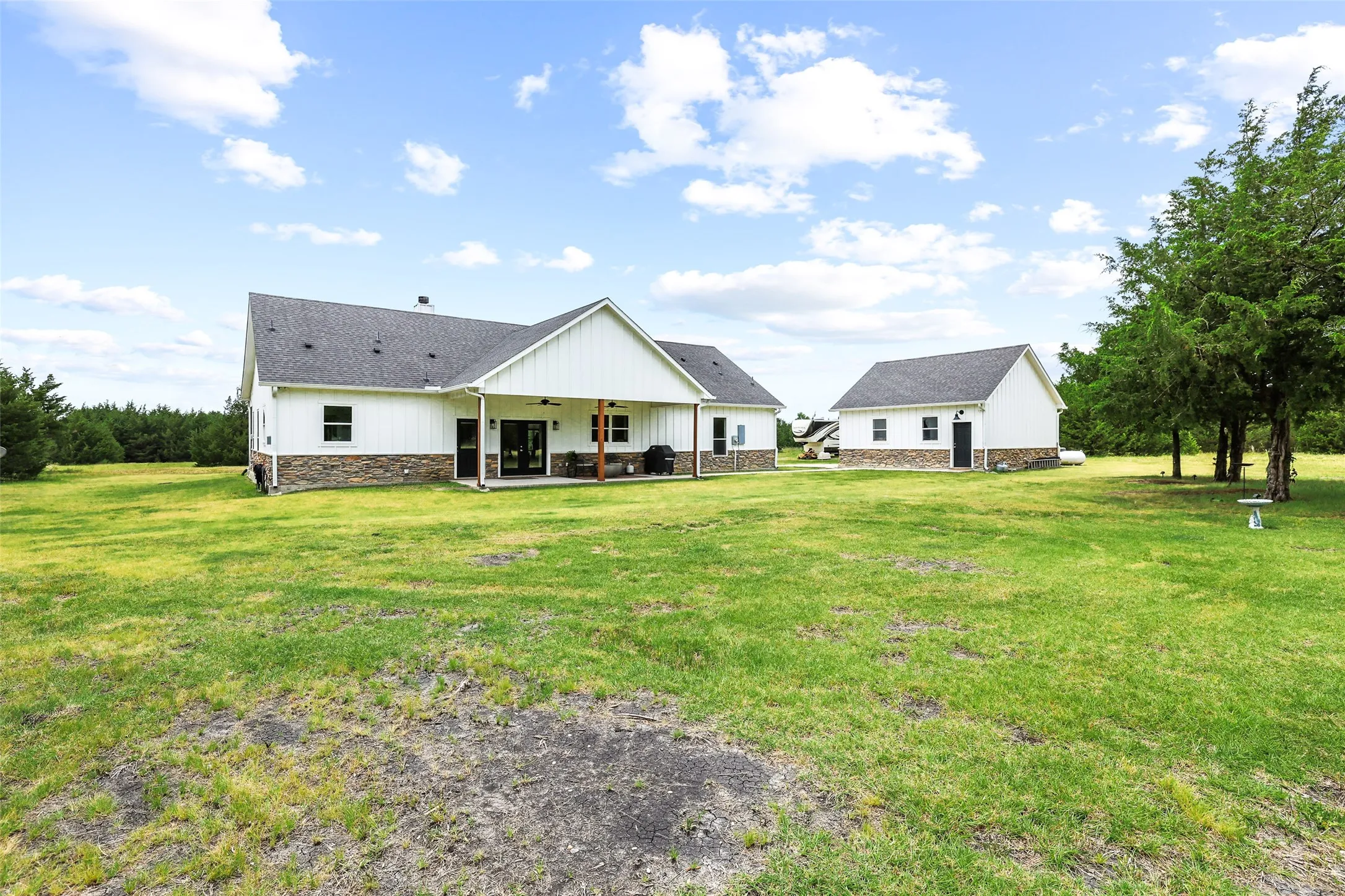 Spacious rear exterior featuring a covered patio with exposed wood beams, a secondary detached structure, white board-and-batten siding, and stone veneer accents