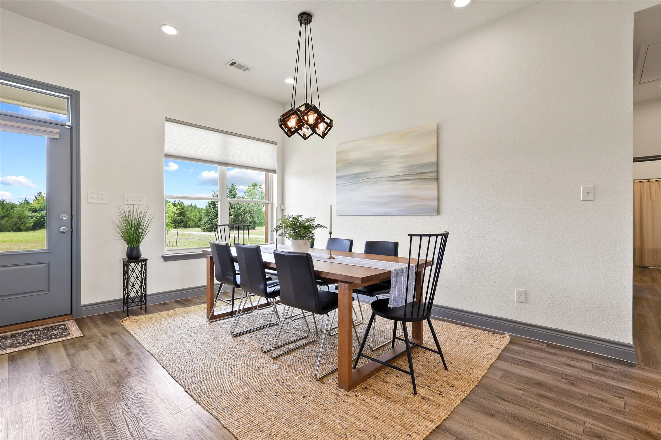 Spacious dining area featuring wood-finish flooring, a contemporary chandelier, large windows with roller shades, and a glass-paneled exterior door