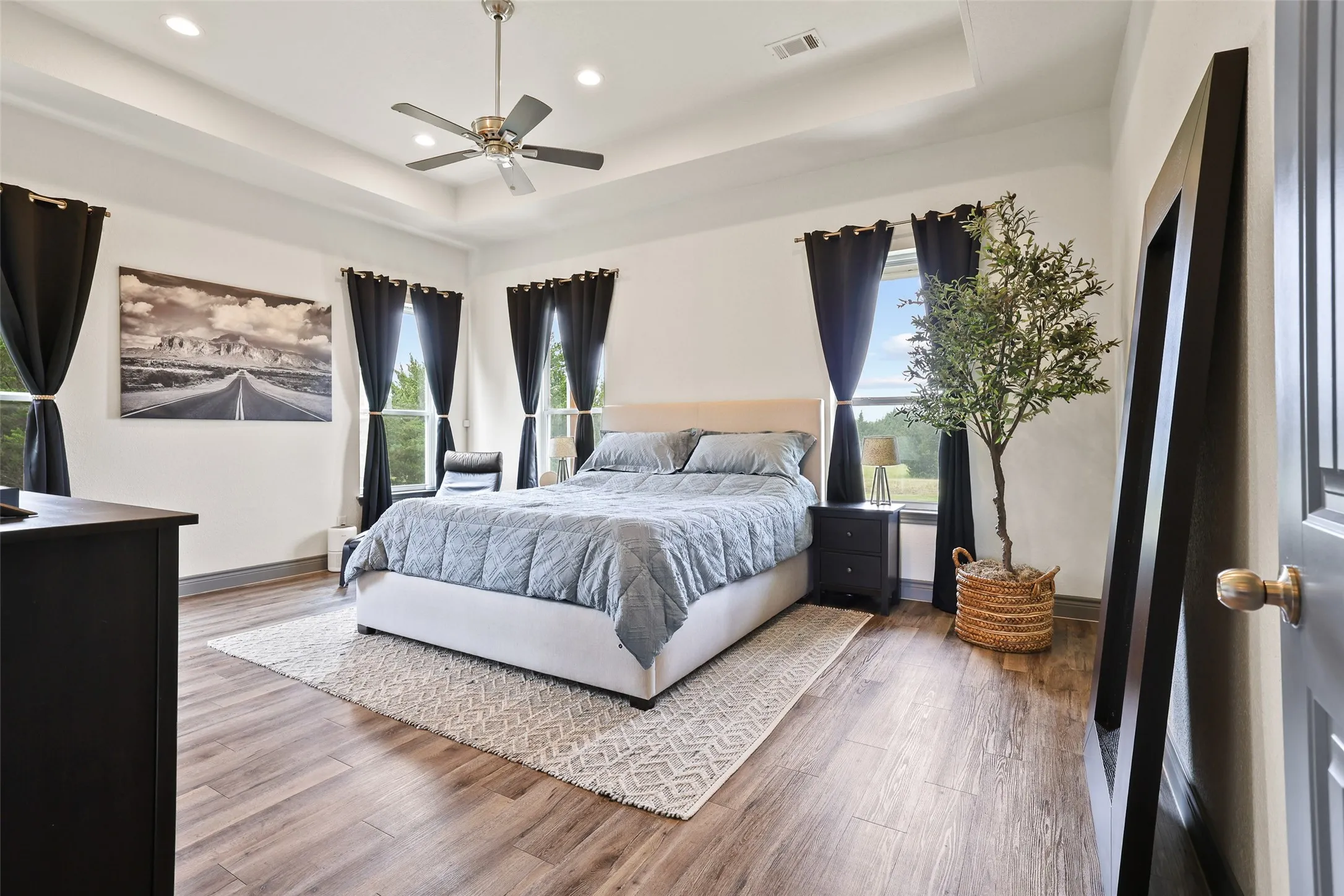 Spacious bedroom featuring wood-finish flooring, a tray ceiling with a ceiling fan, recessed lighting, and multiple windows