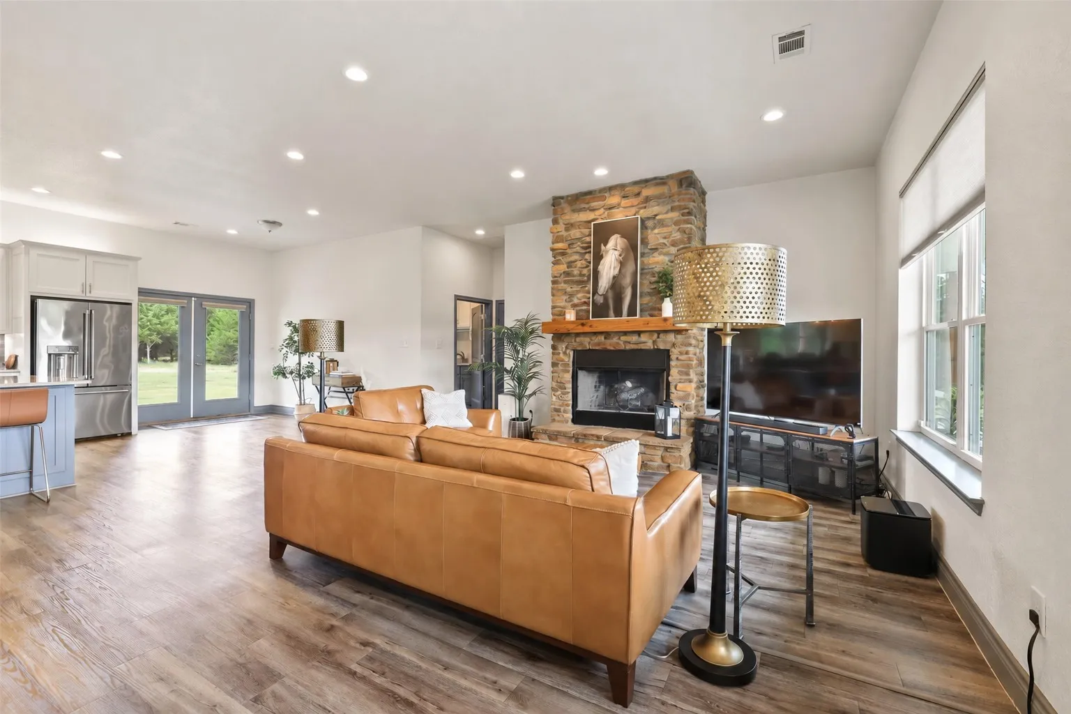 Wood-finish flooring throughout, recessed ceiling lighting, stone fireplace with wood mantel, stainless steel refrigerator, and double French doors to the exterior