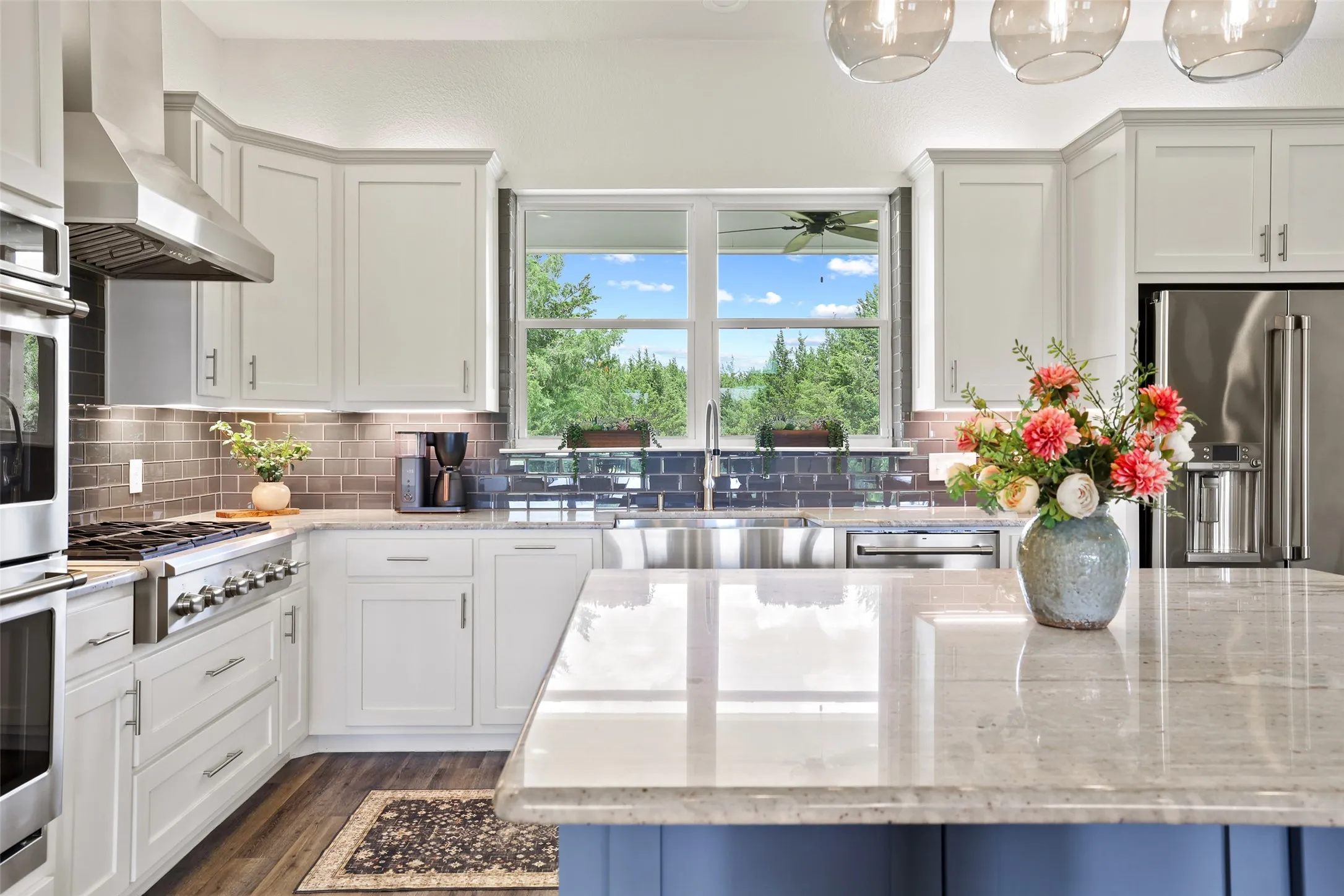 Modern kitchen featuring an expansive island with a light-toned countertop, white cabinetry, a stainless steel range hood, built-in double ovens, and a large window above a stainless steel farmhouse sink