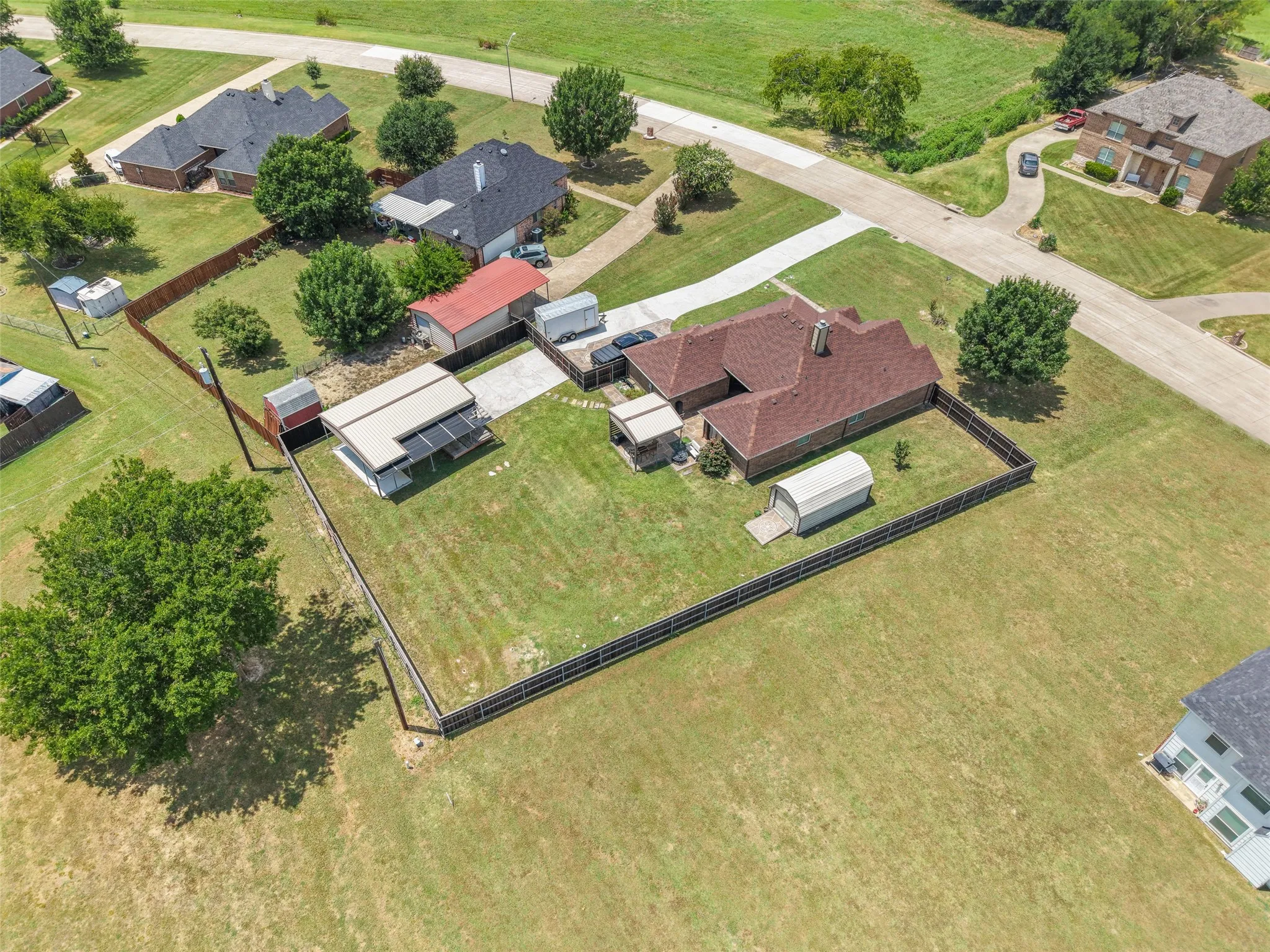 Expansive fenced property featuring a brick residence with a red shingle roof, a detached carport structure, and two additional storage buildings