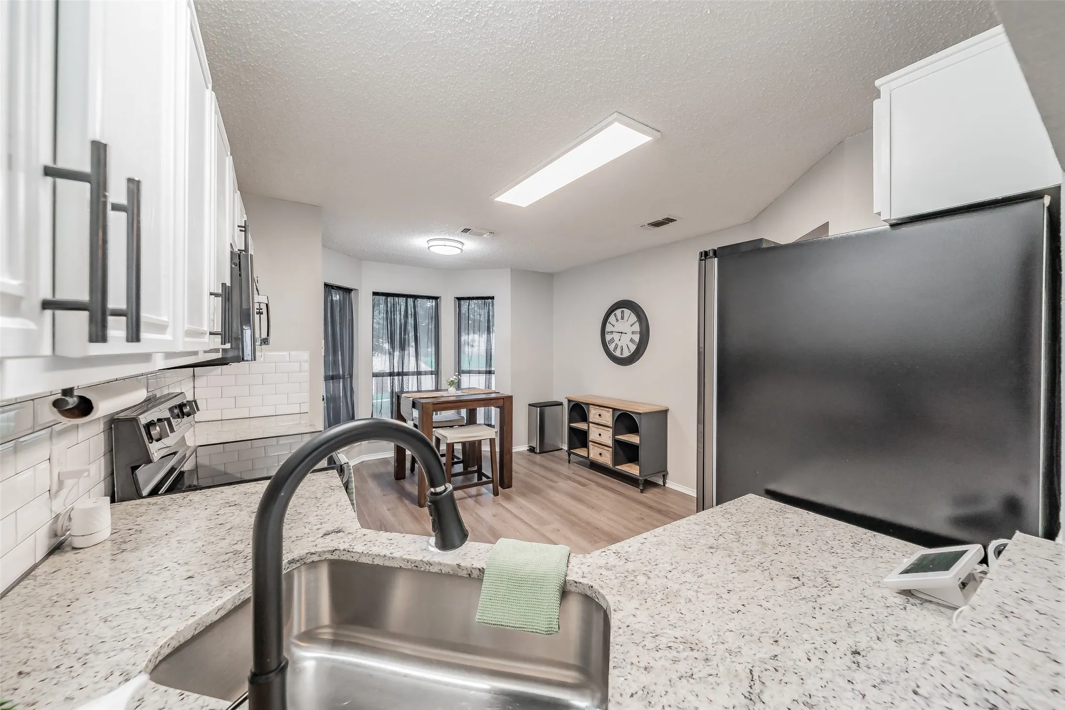 Kitchen featuring a stainless steel sink with a matte black gooseneck faucet, light granite countertops, white subway tile backsplash, and white cabinetry with dark pull hardware