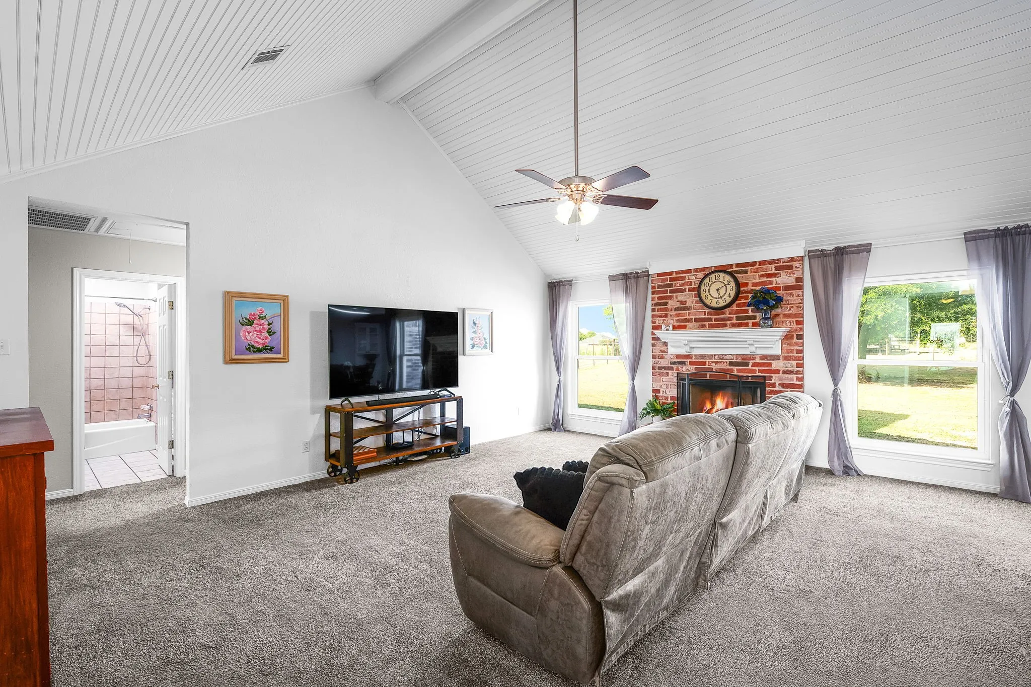 Living area with vaulted white plank ceiling, brick fireplace surround, white mantel, and two large windows