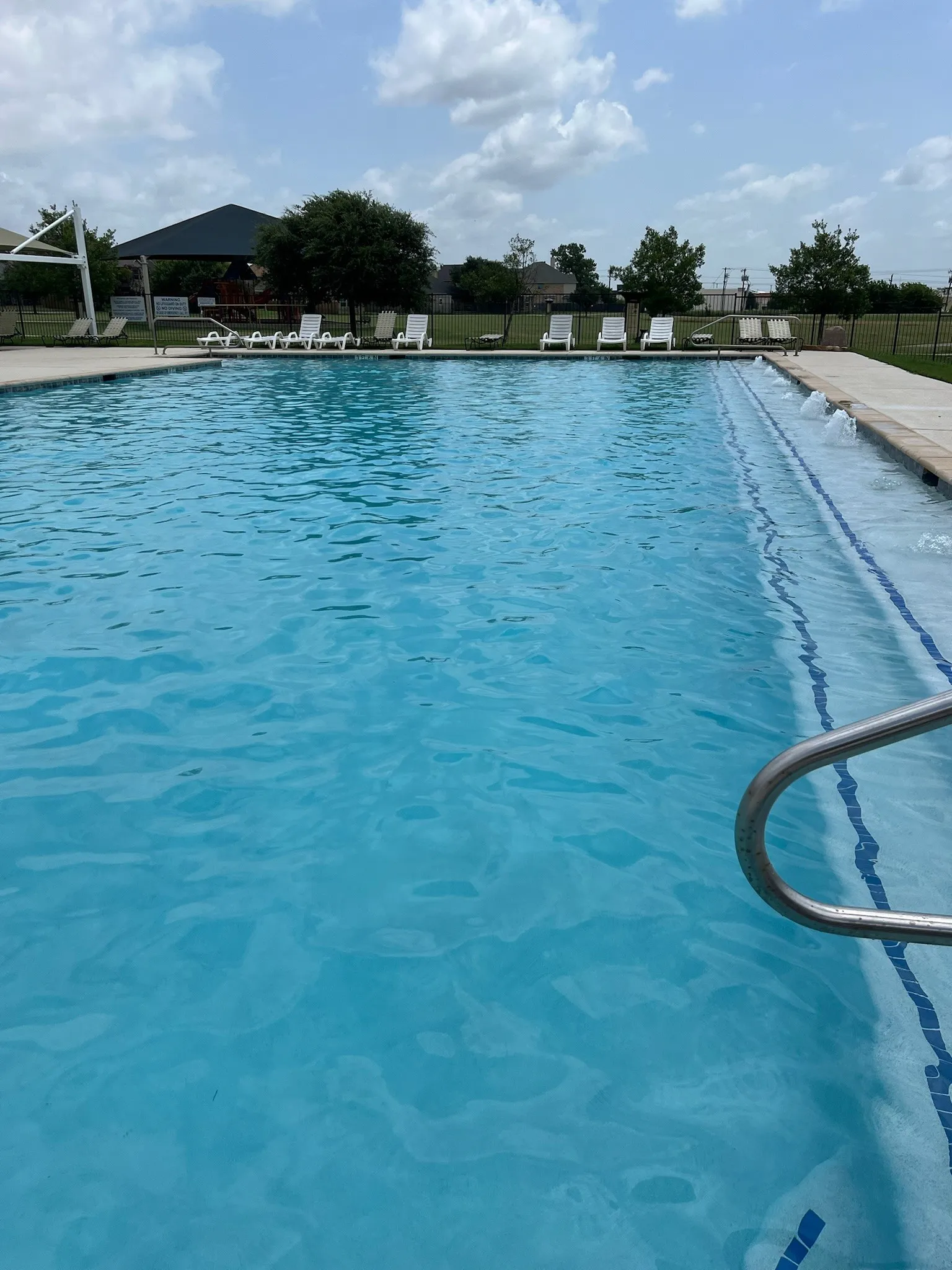 Outdoor swimming pool featuring a tanning ledge with water features, a metal handrail, and lounge chairs on a surrounding deck