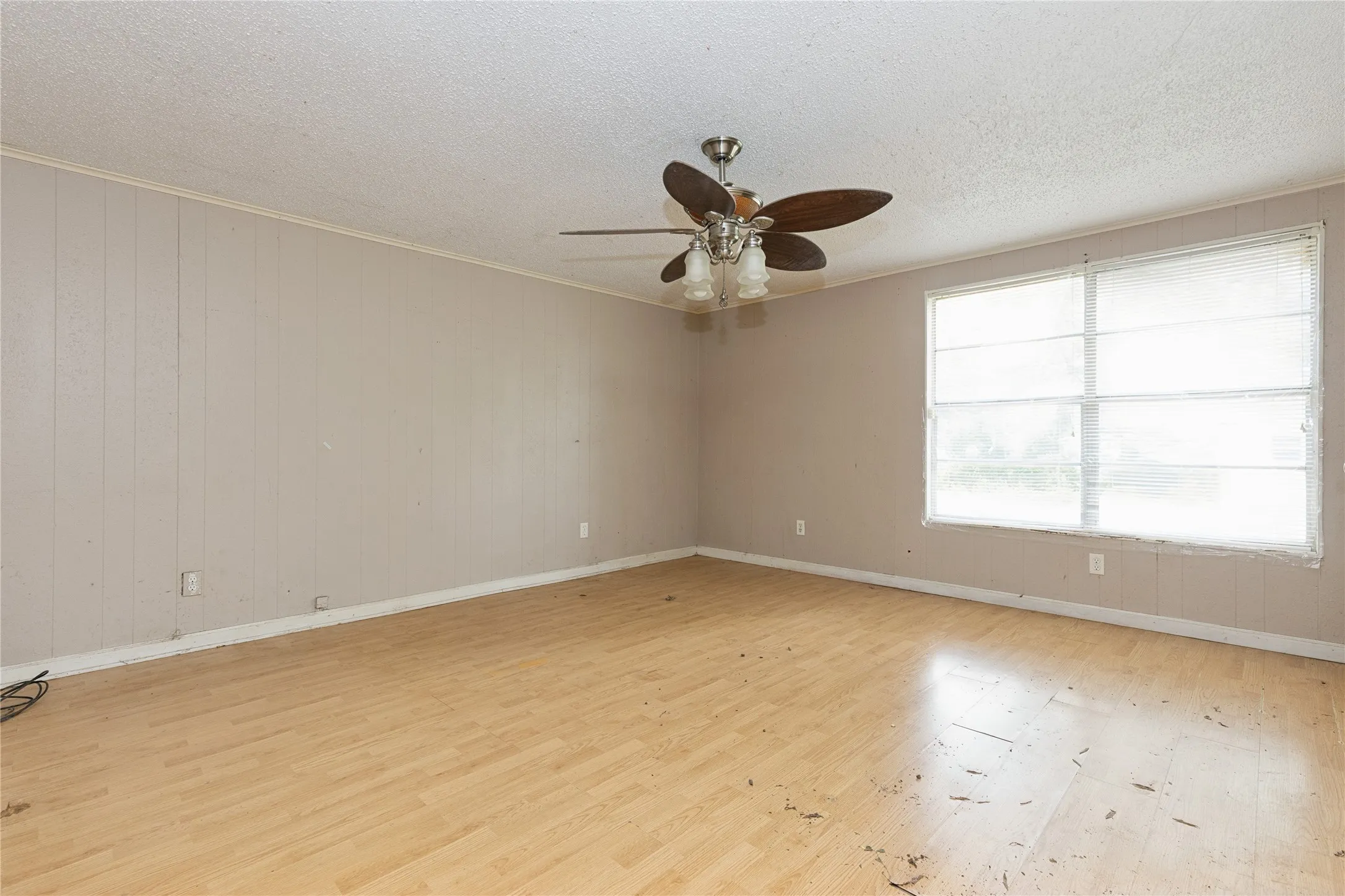 Spacious room featuring light wood-finish flooring, neutral wall paneling, and a large window with blinds