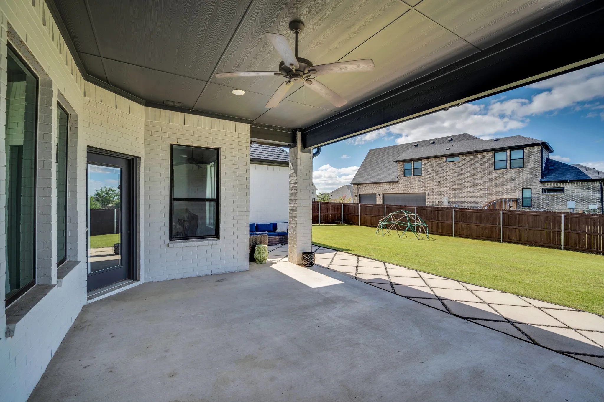 Covered concrete patio with ceiling fan, white brick exterior, black trim windows, and direct access to a manicured lawn