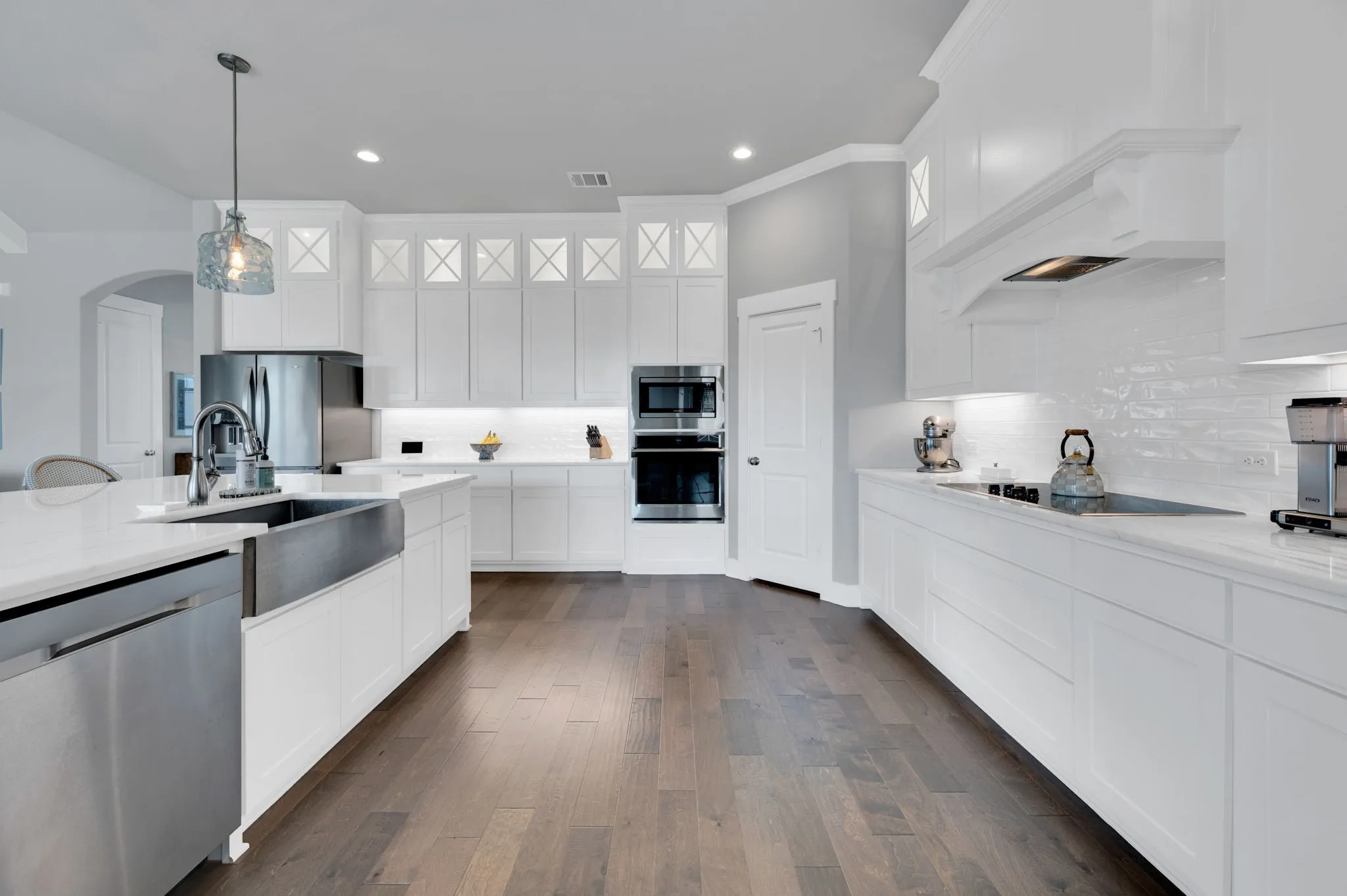 Gourmet kitchen featuring a large island with a stainless steel farmhouse sink, pendant lighting, shaker-style white cabinetry, stainless steel appliances, and wood-finish flooring