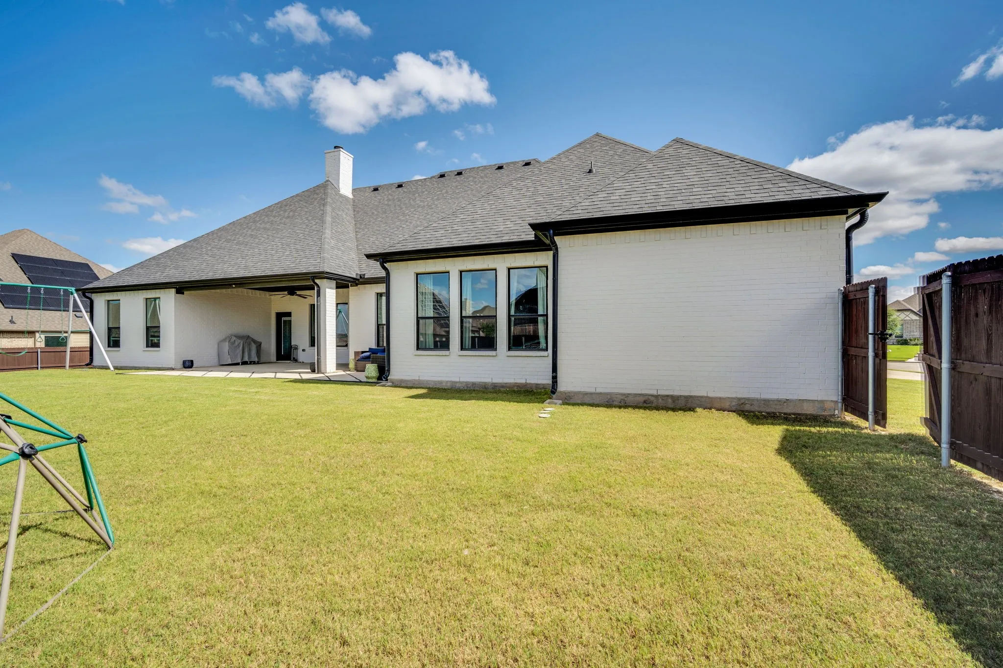 Spacious backyard featuring a covered patio, extensive lawn, white brick exterior, black-framed windows, and a privacy fence with gate