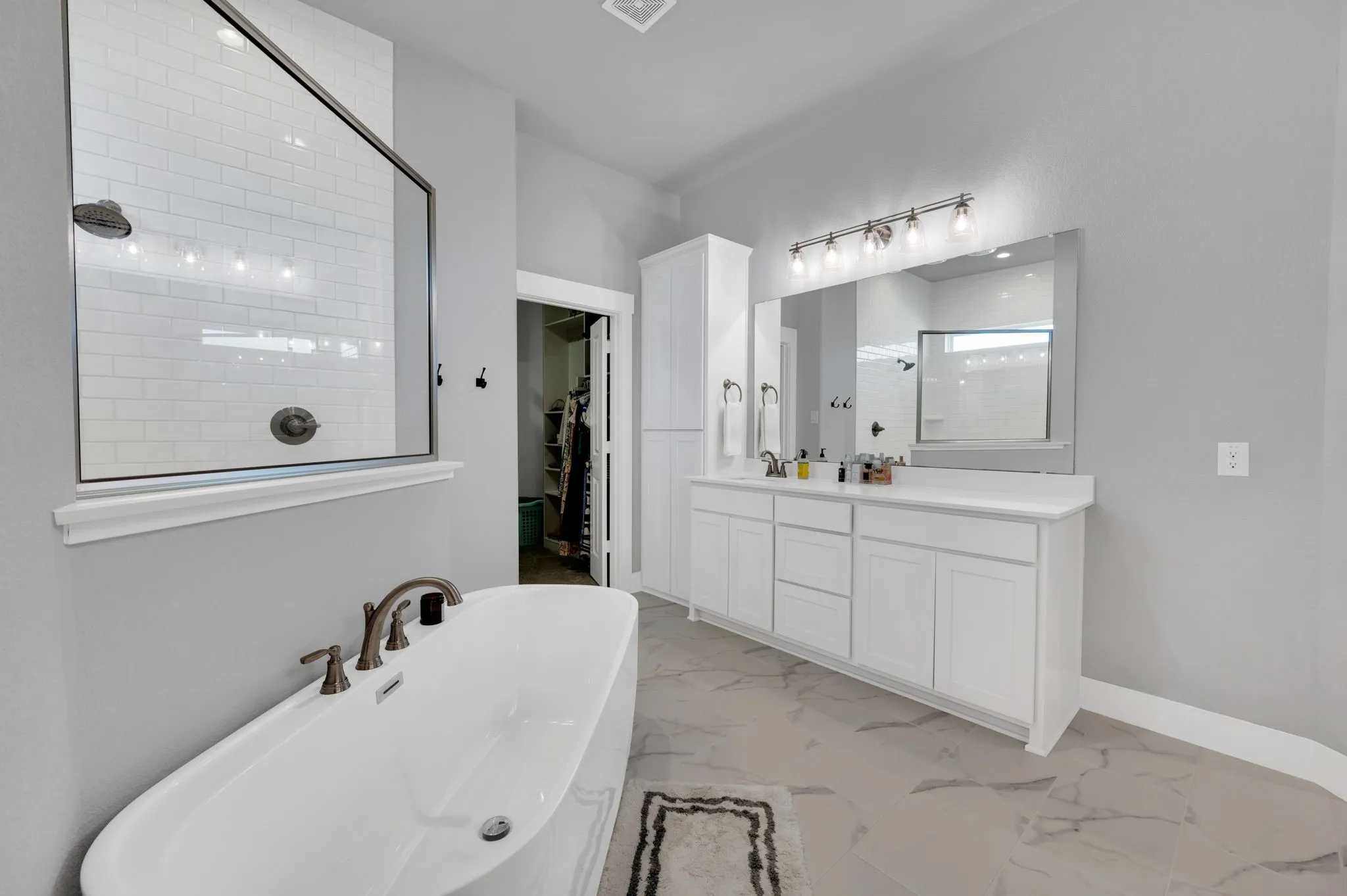Freestanding soaking tub with floor-mounted faucet, white subway tile shower with glass panel, integrated shower shelving, and a dual-sink vanity with quartz countertop