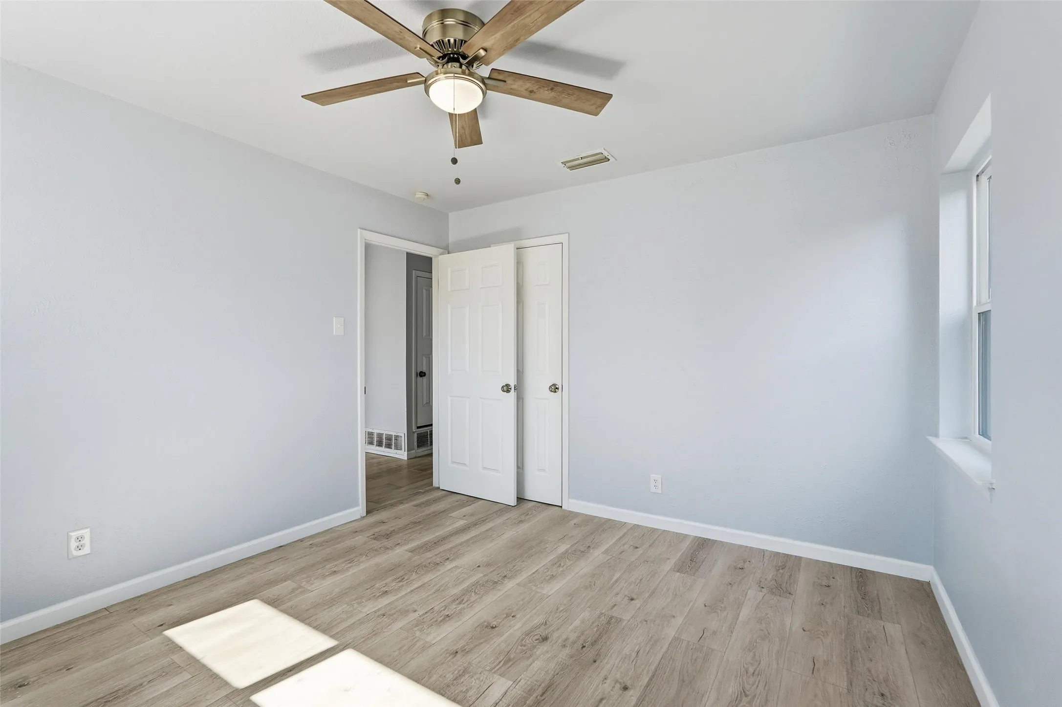 Light-filled room featuring wood-finish flooring, a ceiling fan with integrated lighting, a white trim window, and a paneled door
