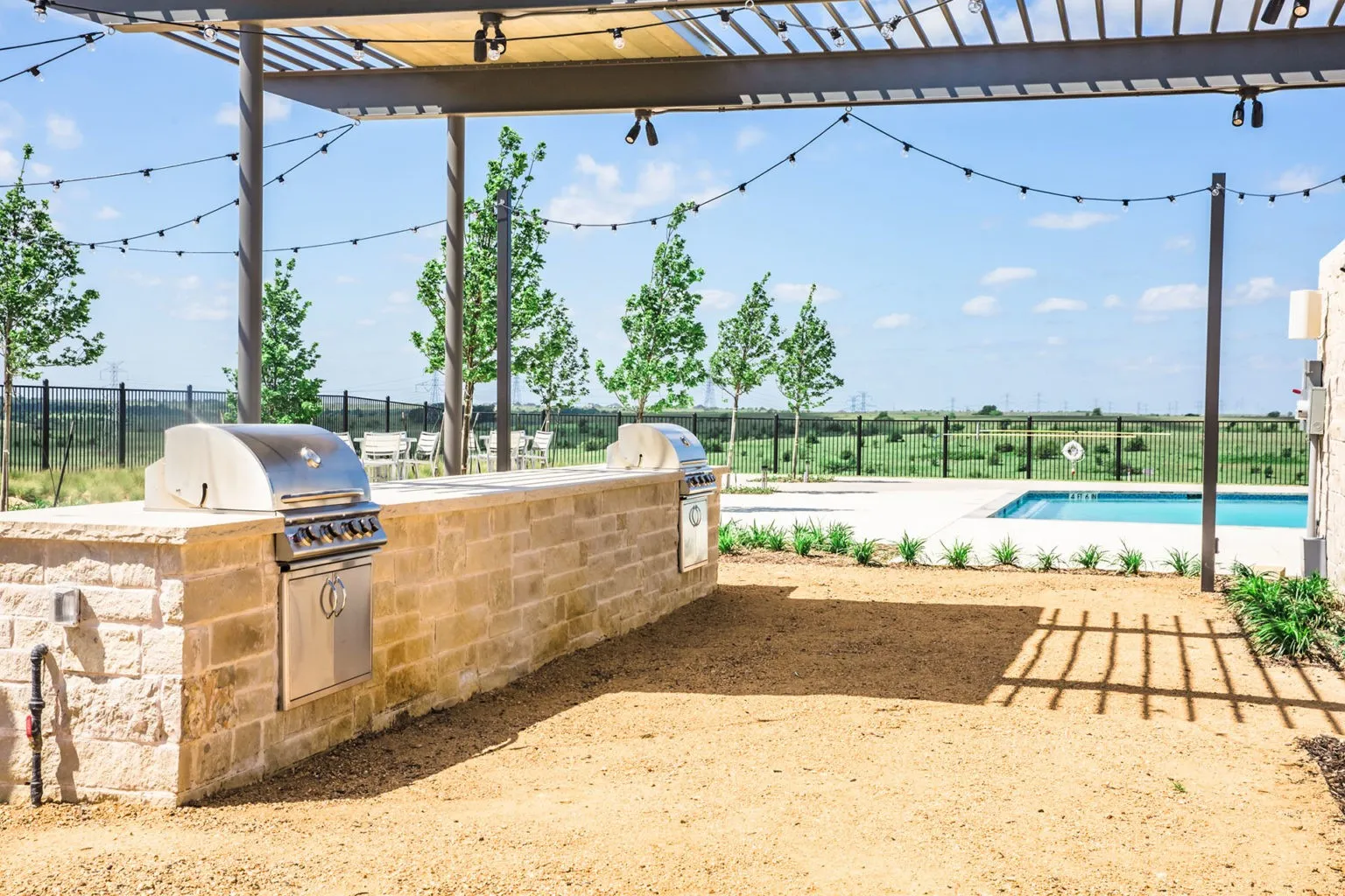 Outdoor entertaining area featuring a stone-clad grill station with two stainless steel grills, a pergola structure, string lighting, and direct access to a swimming pool