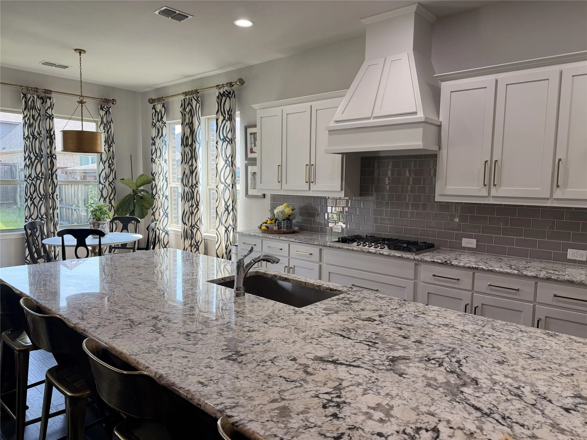 Spacious kitchen featuring an expansive island with a stainless steel undermount sink, white shaker cabinetry with dark hardware, a subway tile backsplash, a built-in gas cooktop, and a custom range hood