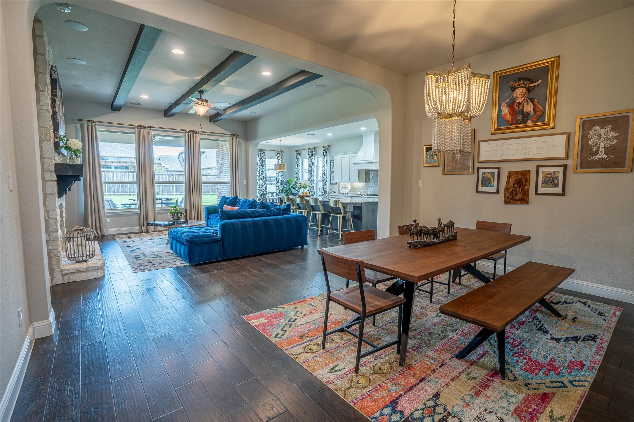 Open-concept interior featuring wood-finish flooring, a dining area with a multi-tiered chandelier, and a living area with exposed beam ceilings and a stone fireplace
