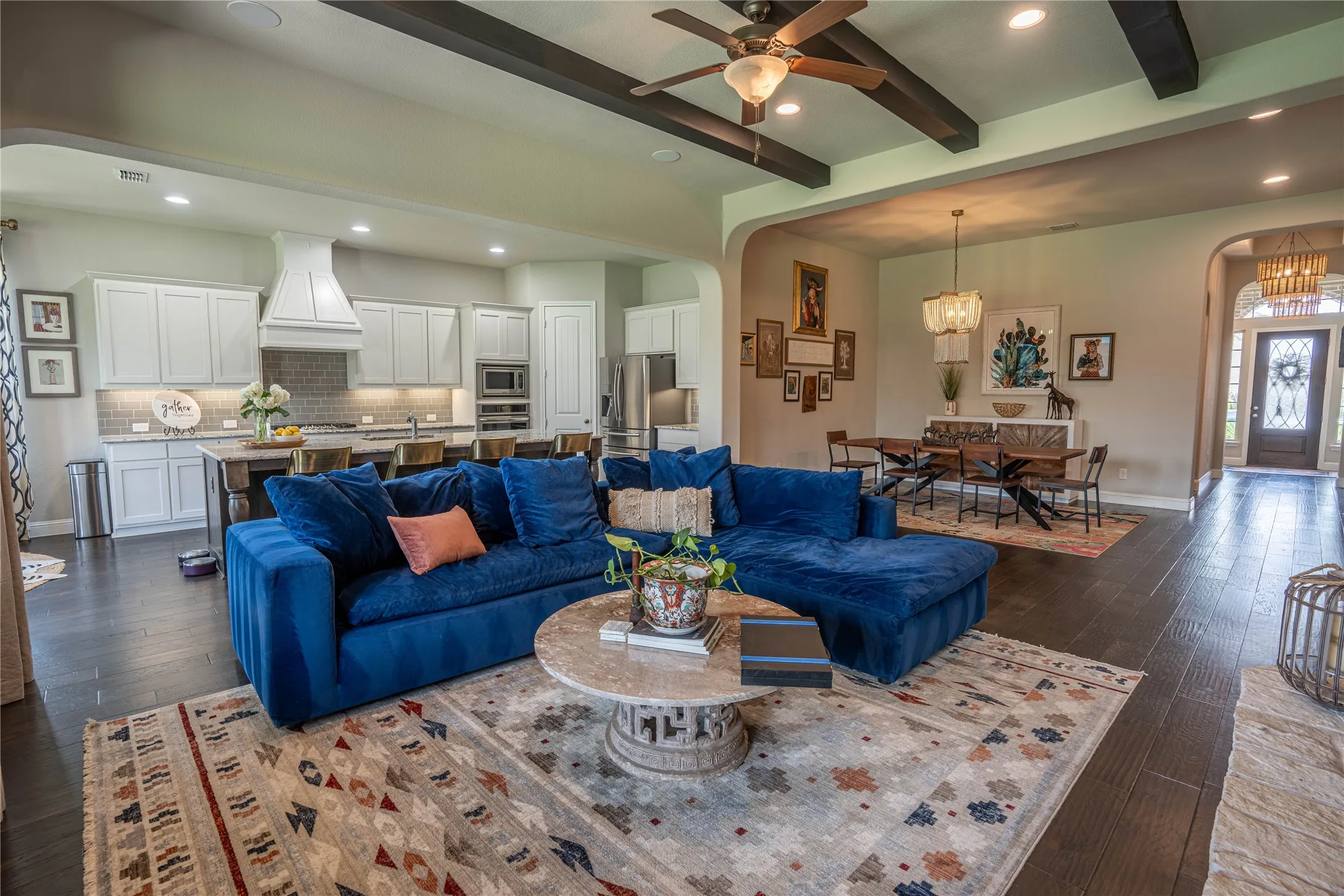 Open-concept living space featuring wood-finish flooring, a kitchen with white cabinetry and an island, a dining area with chandelier lighting, and exposed beam ceiling details
