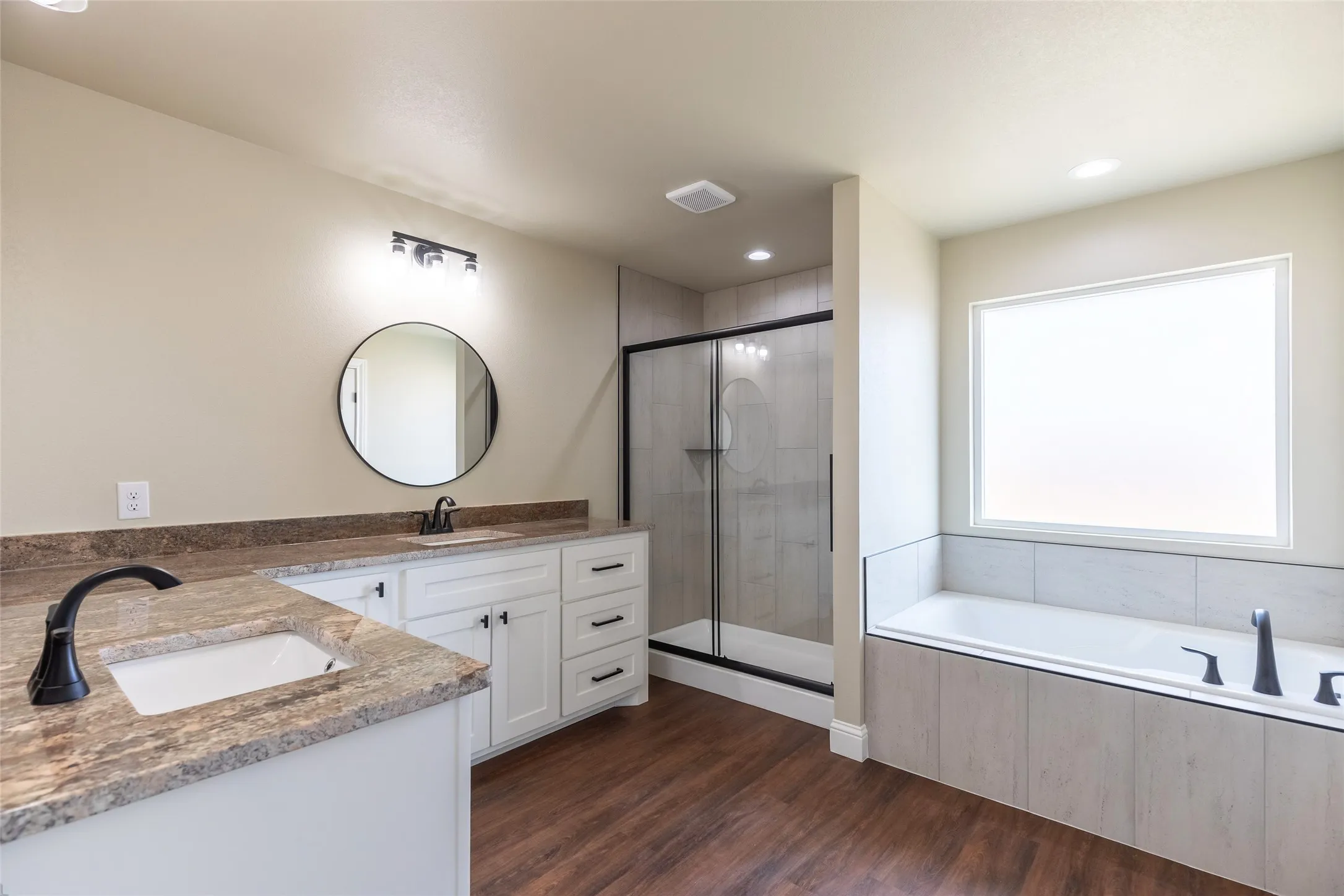 Bathroom featuring a dual vanity with undermount sinks and stone countertops, a soaking tub with tile surround, a walk-in shower with clear glass enclosure, and wood-finish flooring