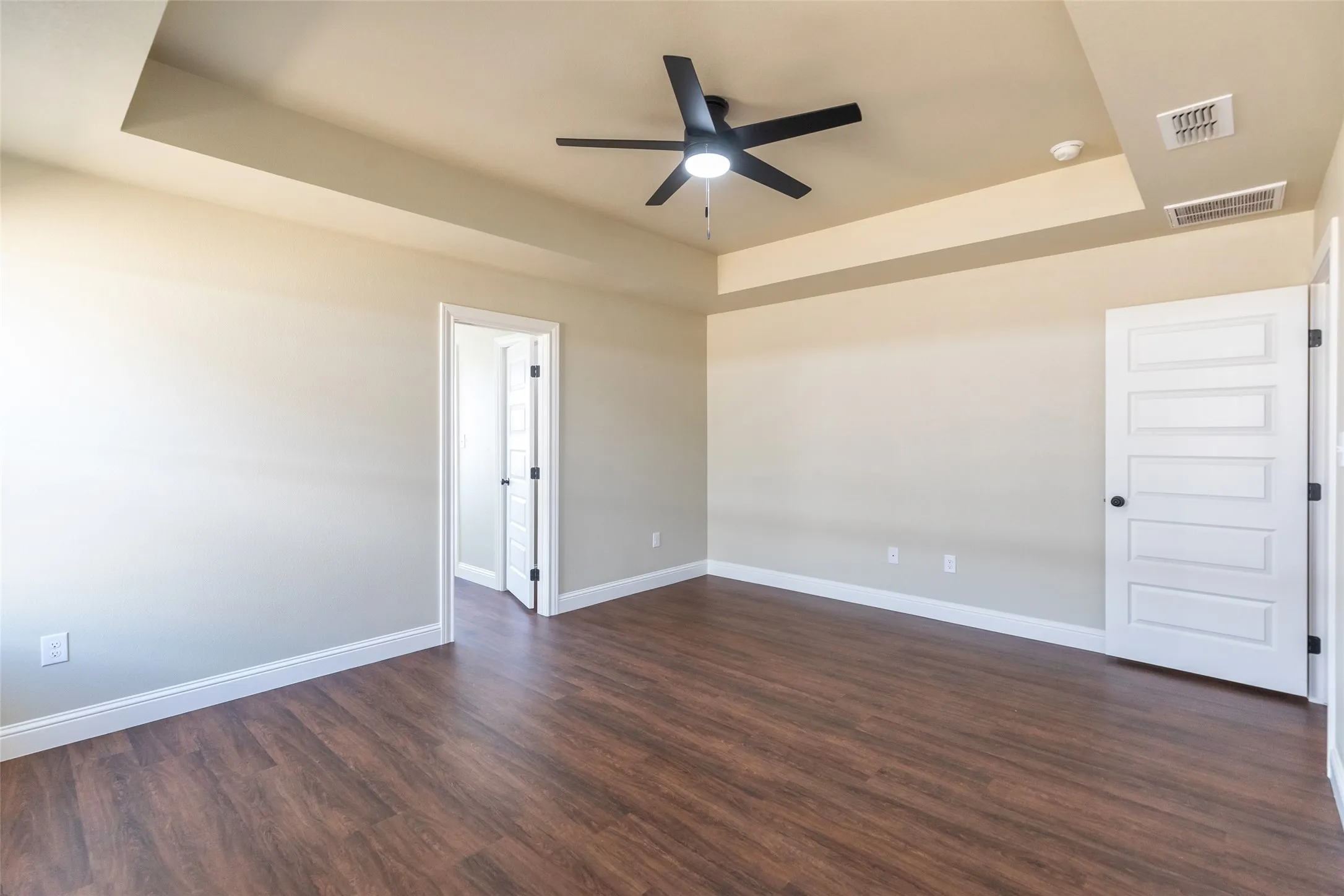 Spacious room featuring wood-finish flooring, light neutral wall tones, and a tray ceiling with a black ceiling fan