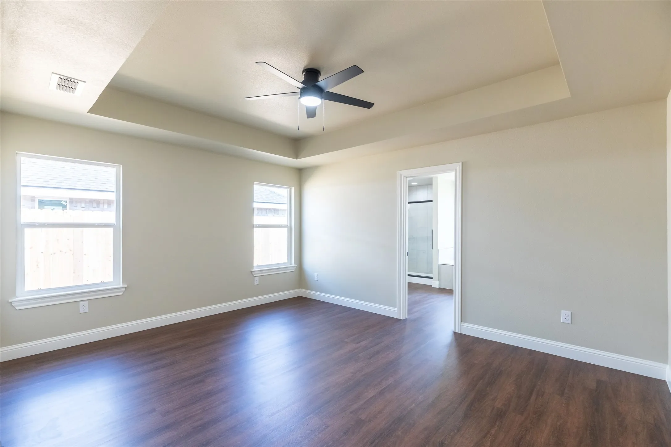 Spacious room featuring wood-finish flooring, a tray ceiling with a ceiling fan, dual windows, and an en-suite bathroom entry