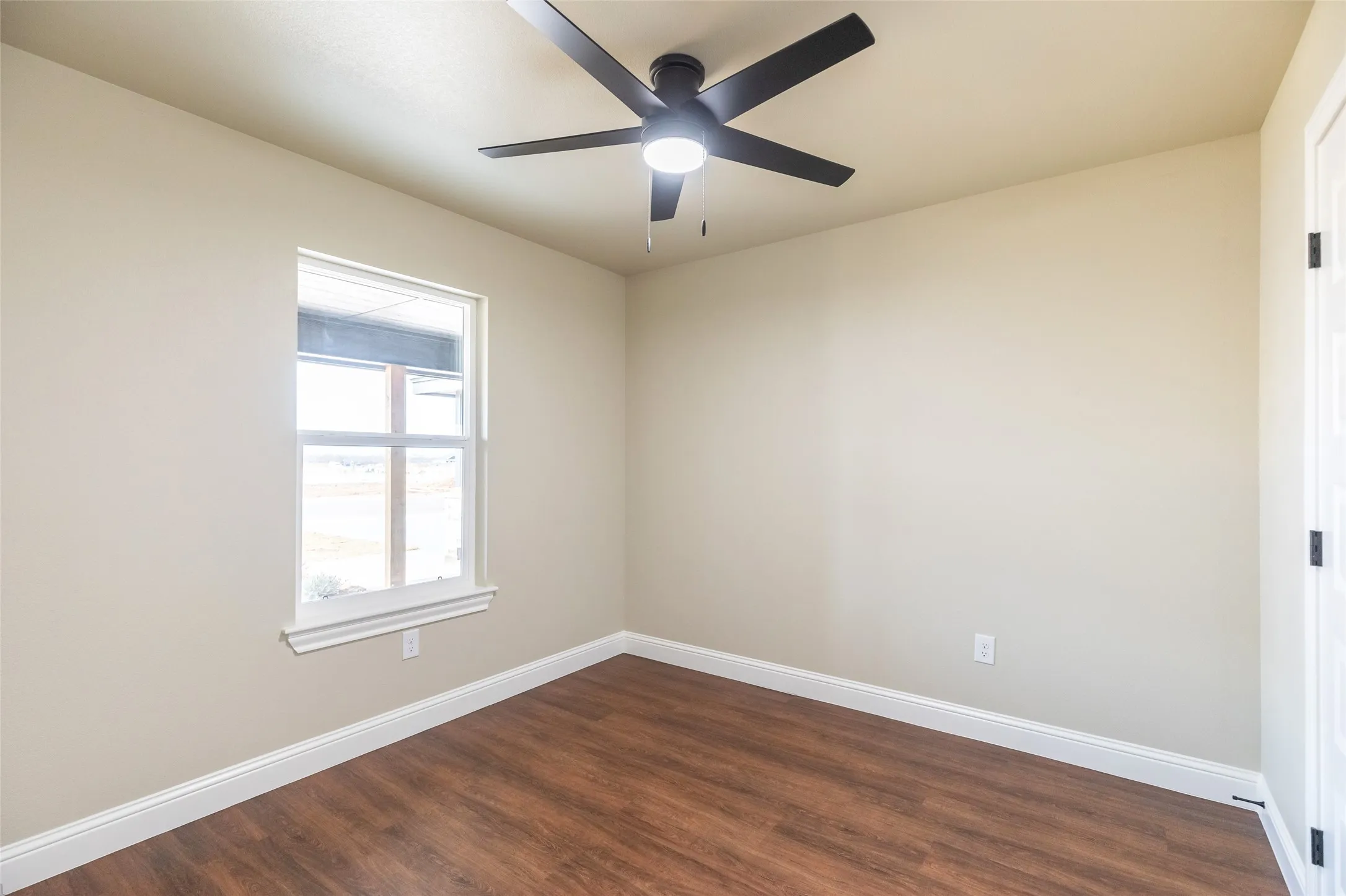 Neutral-toned interior room featuring wood-finish flooring, a white-framed window, and a ceiling fan with integrated lighting