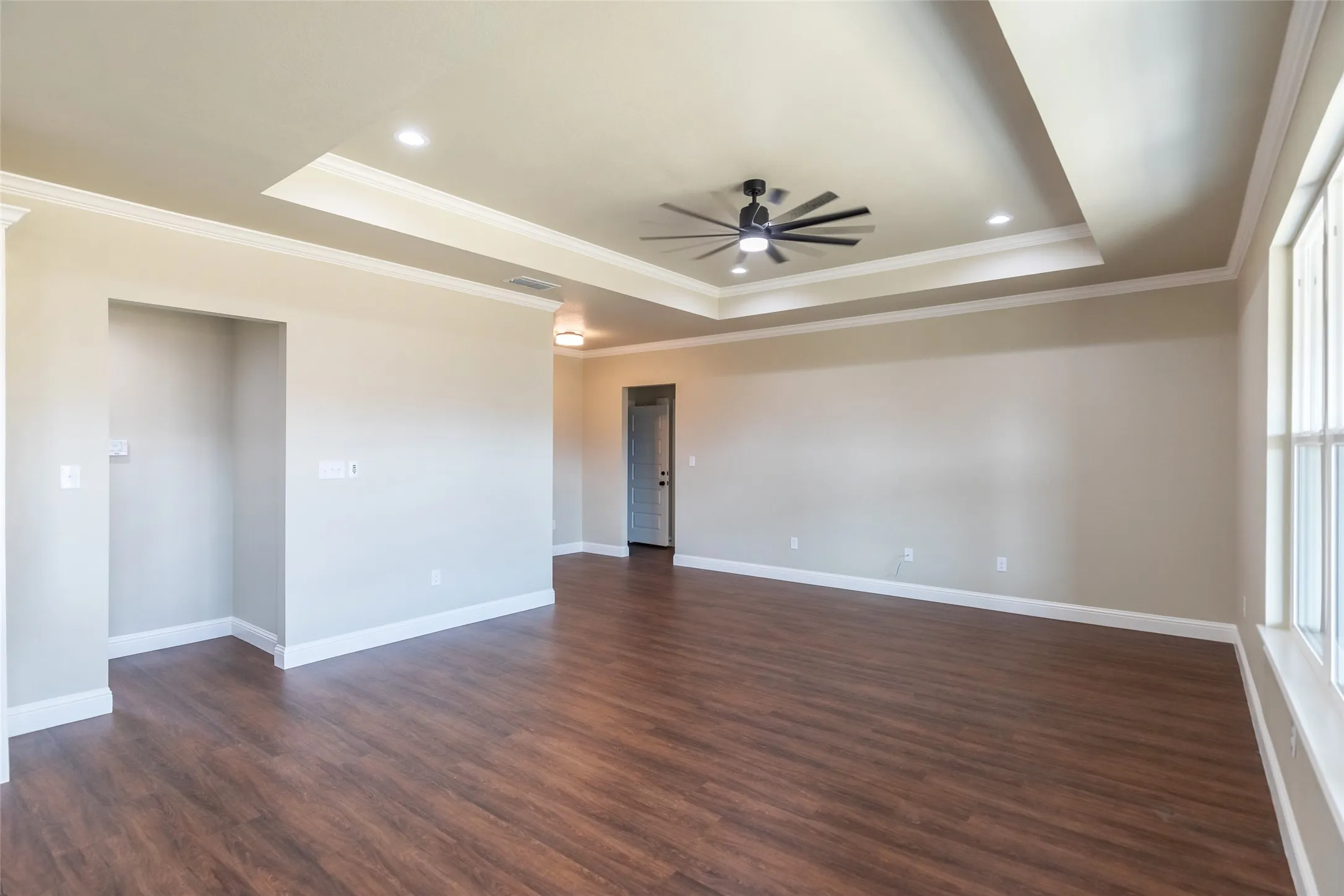 Spacious room with wood-finish flooring, a tray ceiling with crown molding, a ceiling fan, recessed lighting, and large windows