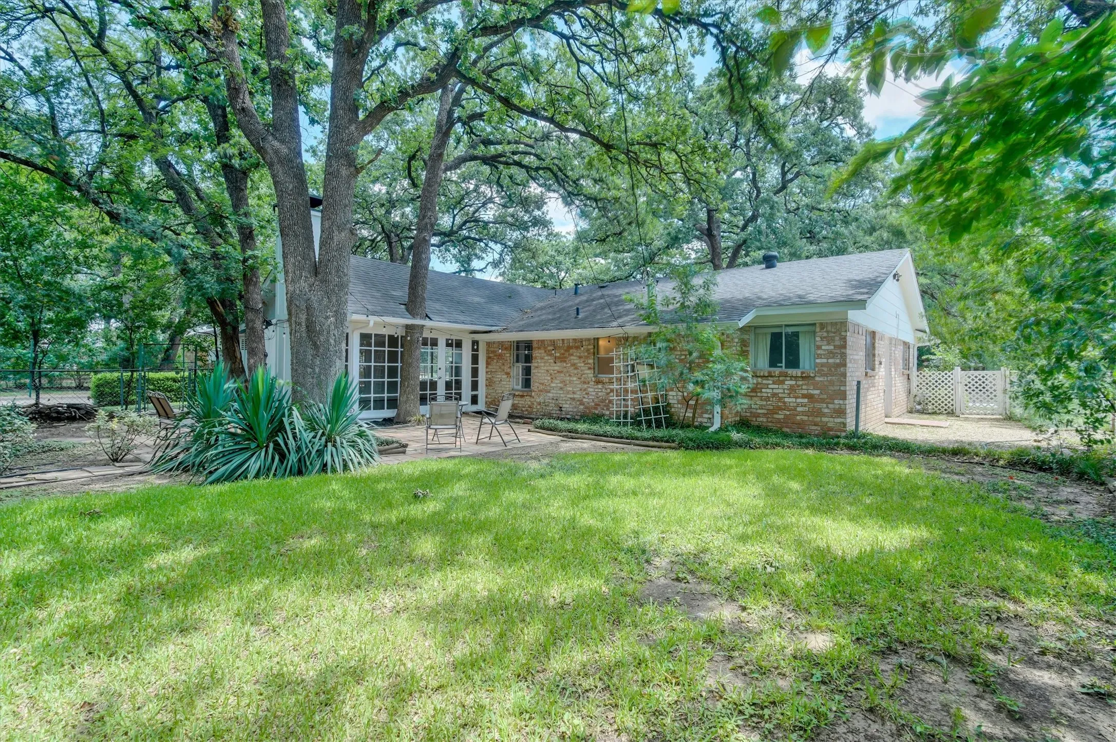 Expansive backyard featuring a large green lawn, mature trees, and a brick exterior home with a patio area