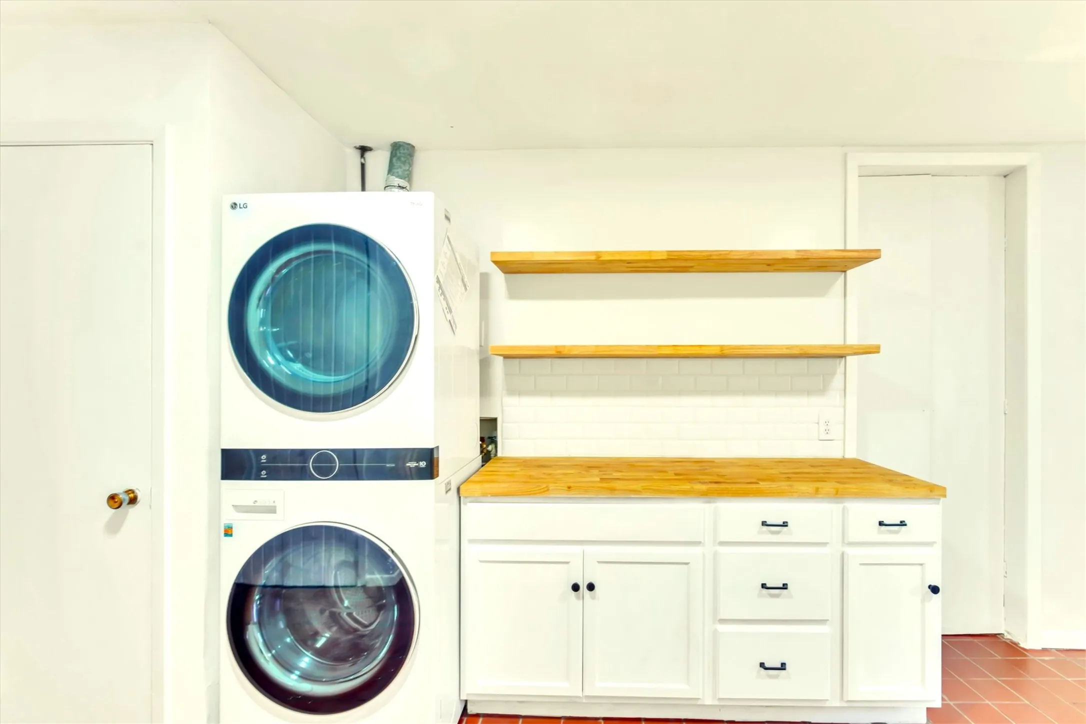Laundry area featuring a stacked washer and dryer, white cabinetry with black hardware, wood-finish countertops, and matching open shelving over a white subway tile backsplash