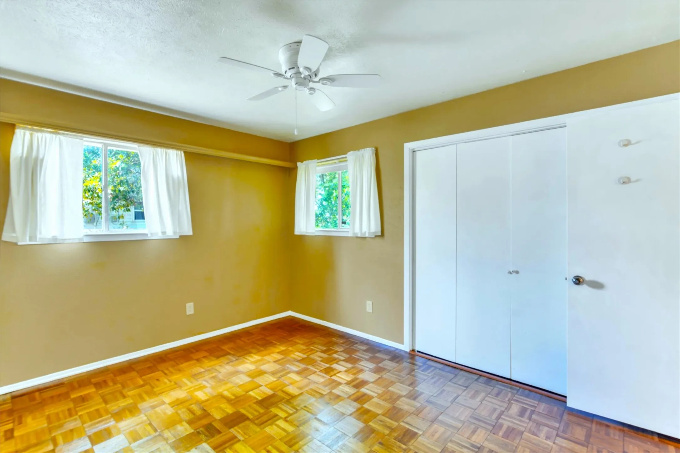 Room featuring parquet flooring, two windows with white trim, a ceiling fan, and a bi-fold closet